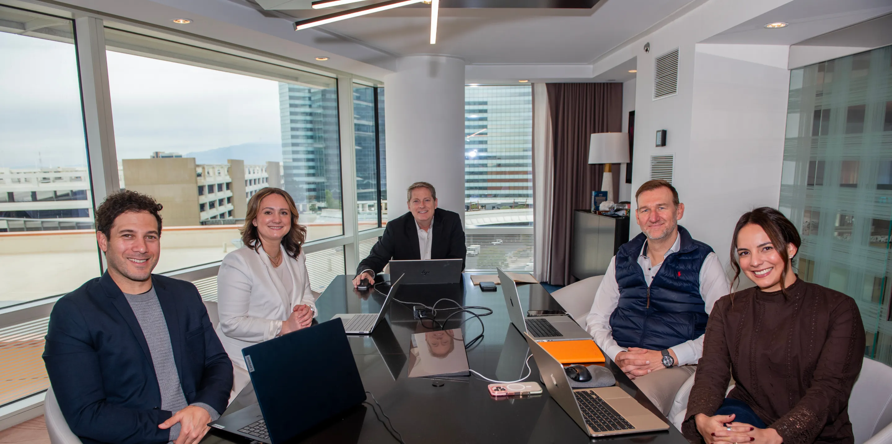 Five businesspeople sitting around a table with laptops in a modern office with city views.