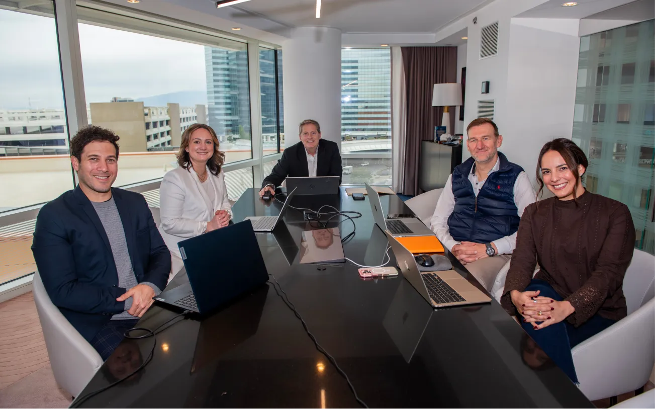 Five diverse professionals smiling around a conference table with laptops in a glass-walled office.