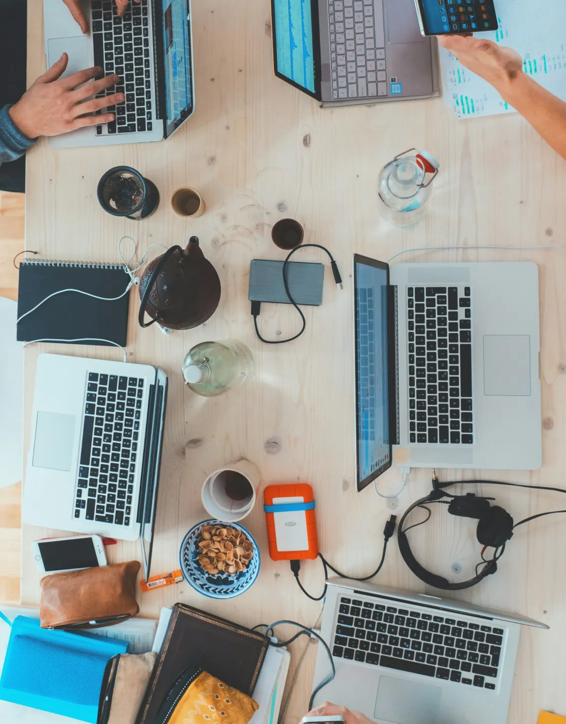 Overhead view of a wooden table with laptops, notebooks, phones, headphones, and snacks with people working.