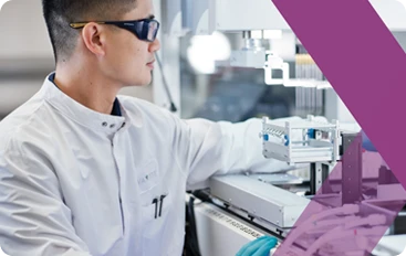 Scientist in white coat and safety glasses working with laboratory automation equipment.