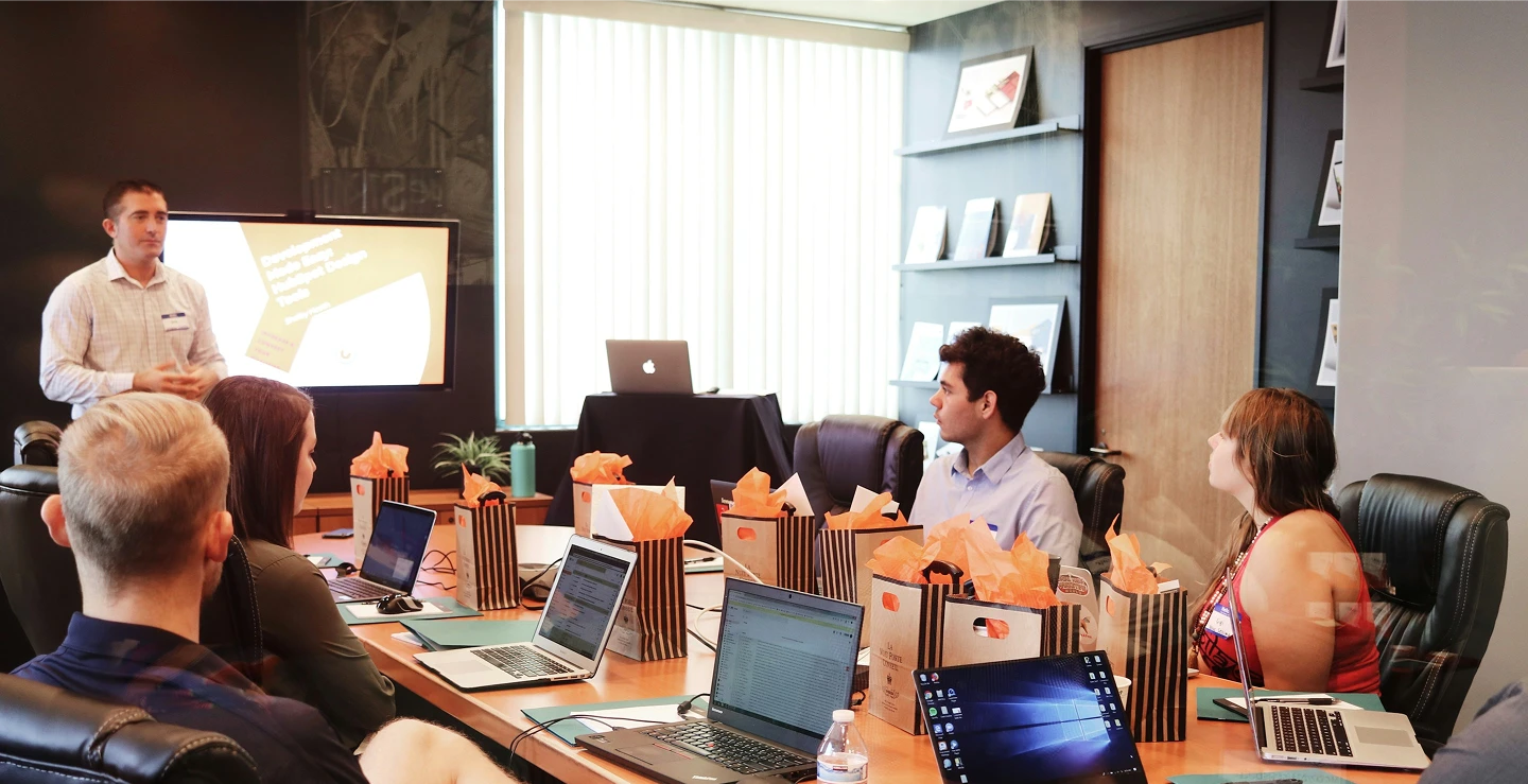 Business meeting with a presenter and five attendees seated at a table with laptops and gift bags.