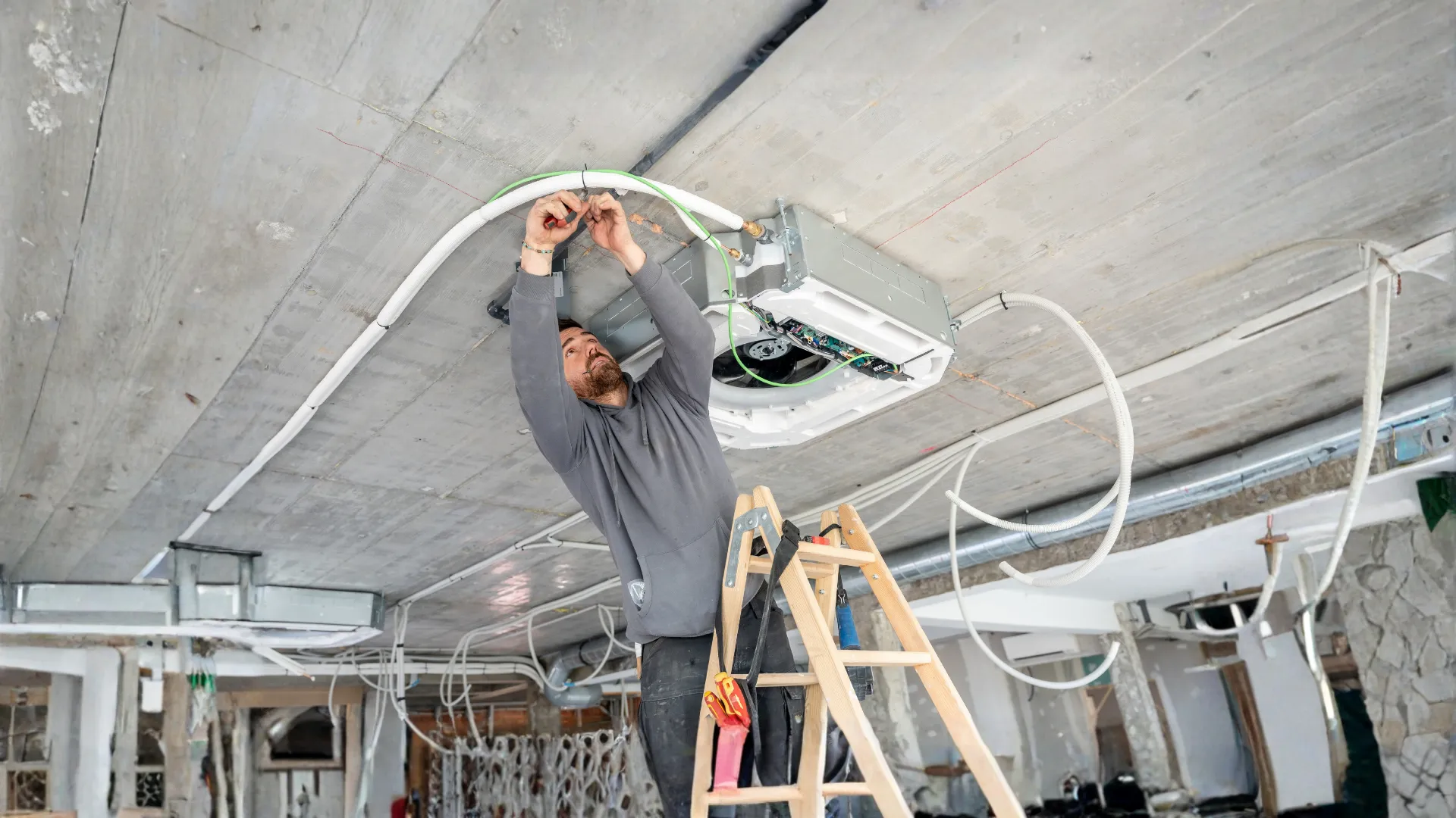 Worker installing HVAC unit on ceiling with wiring on ladder in unfinished building.