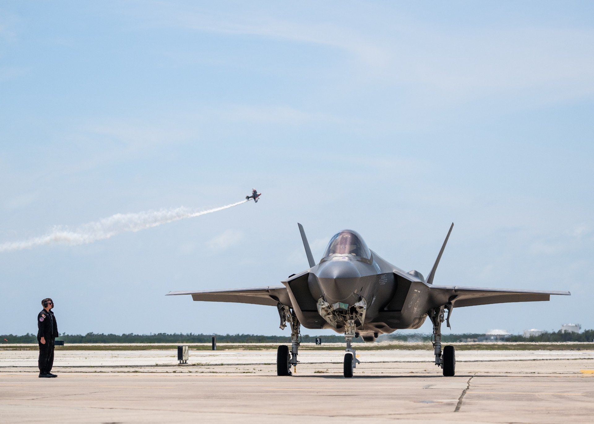 Front view of a fighter jet on a runway with a pilot standing nearby and another aircraft flying overhead leaving a smoke trail.