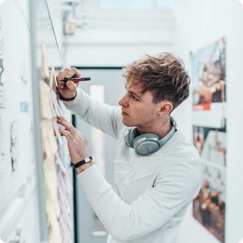 Young man in white hoodie writing on sticky notes on a whiteboard with headphones around his neck.
