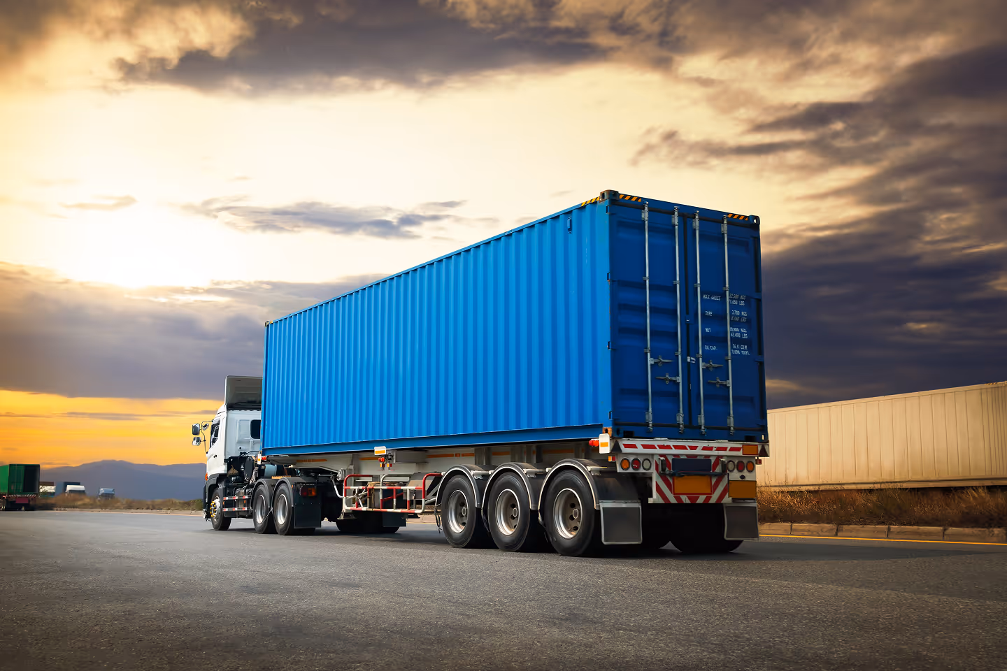 Semi truck with a bright blue shipping container driving on a road at sunset with a cloudy sky.