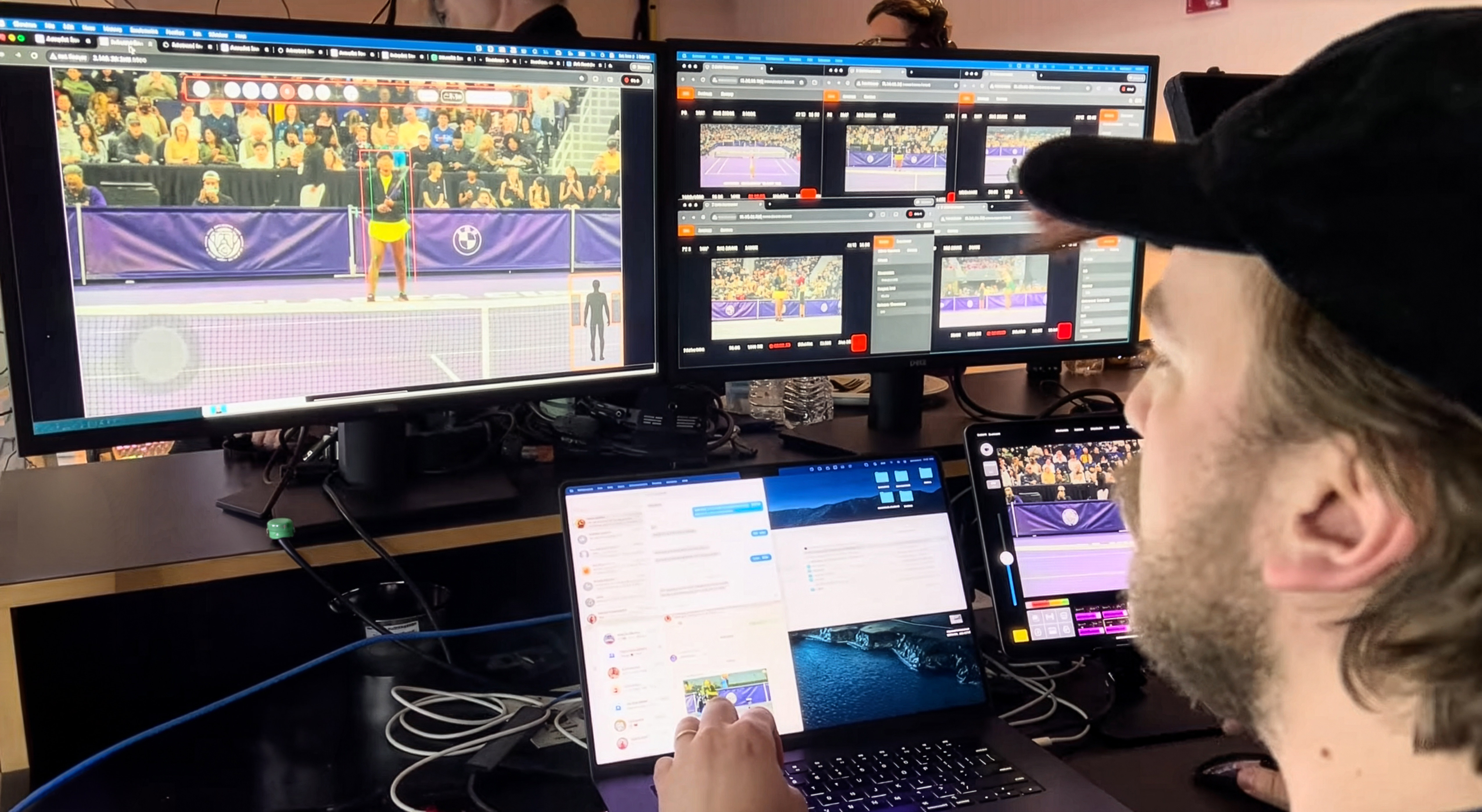 Person wearing a black cap using a laptop and a tablet at a desk with two large monitors displaying tennis match footage and analysis software with multiple camera angles and player tracking.