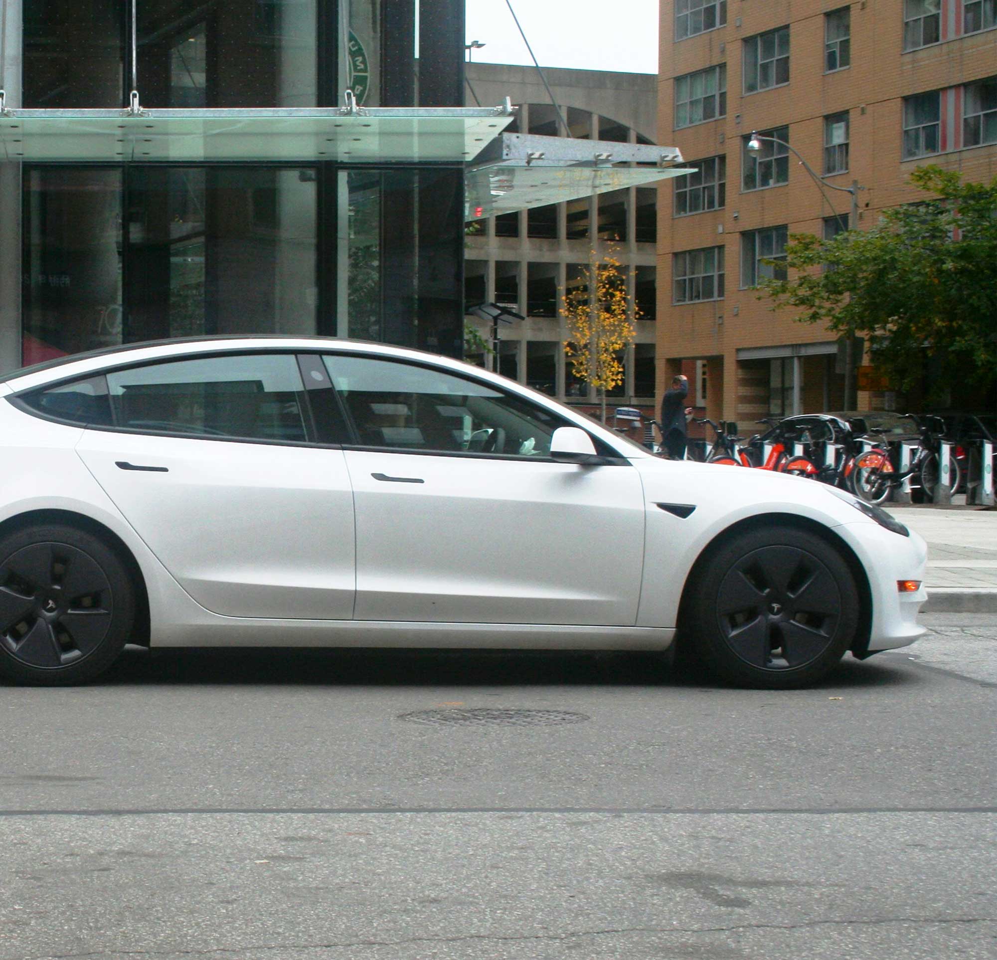 White Tesla electric sedan parked on a city street with a bicycle sharing station and buildings in the background.
