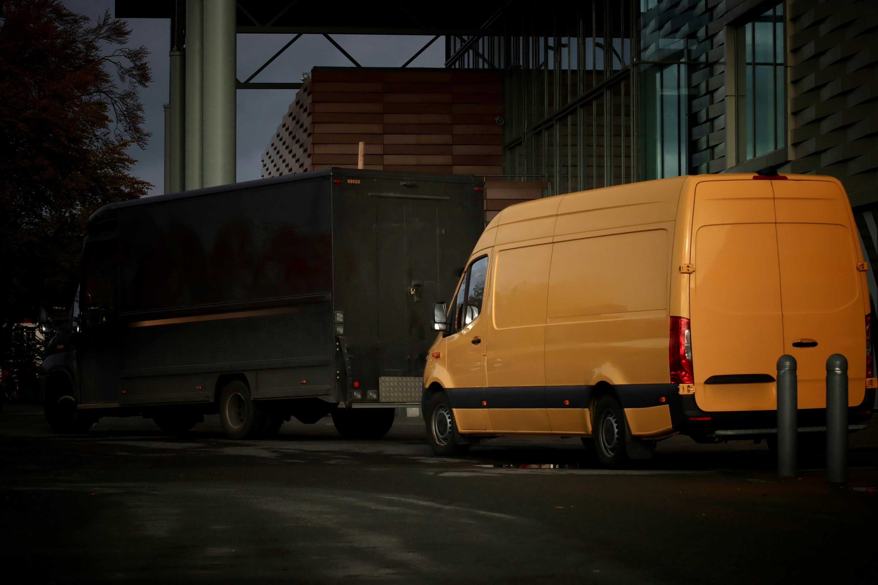 Yellow delivery van parked next to a dark truck beside a modern building.