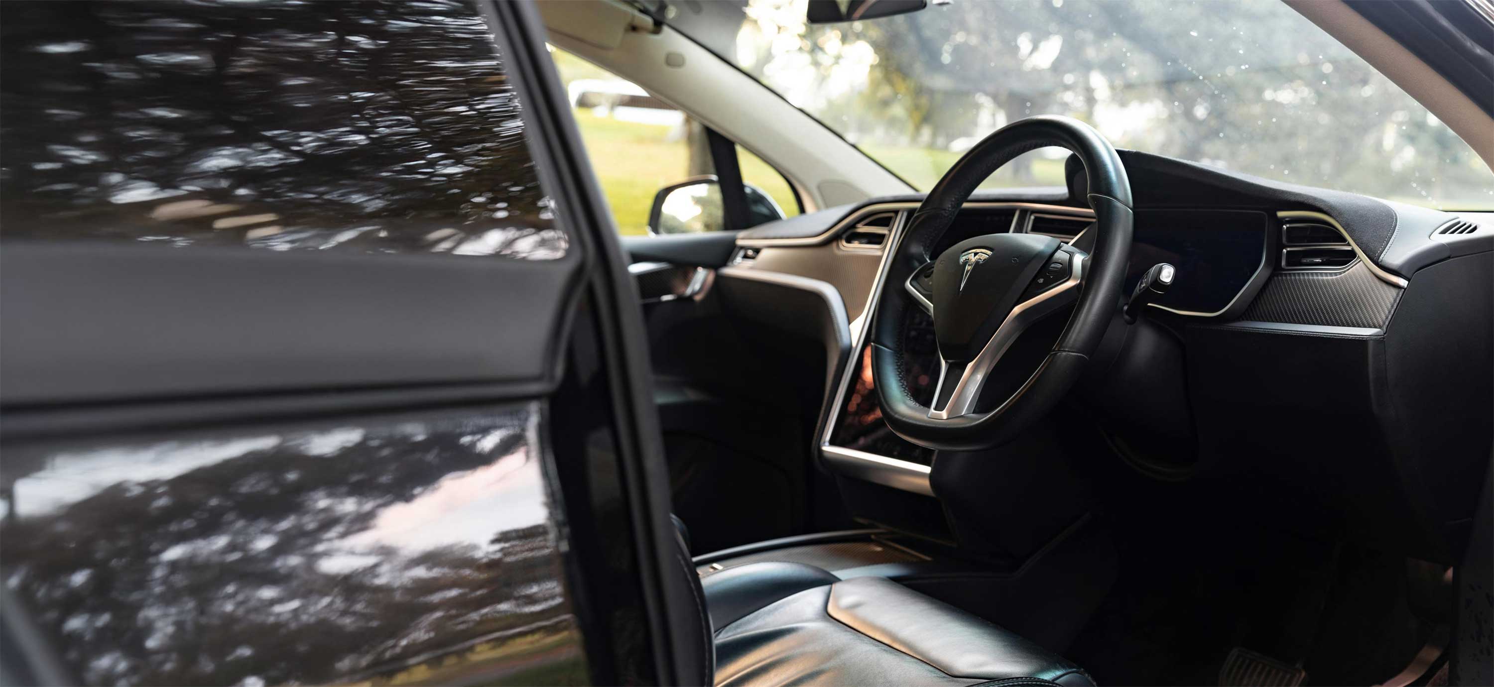 Interior of a Tesla car showing black leather seats, steering wheel with Tesla logo, and dashboard with touchscreen.