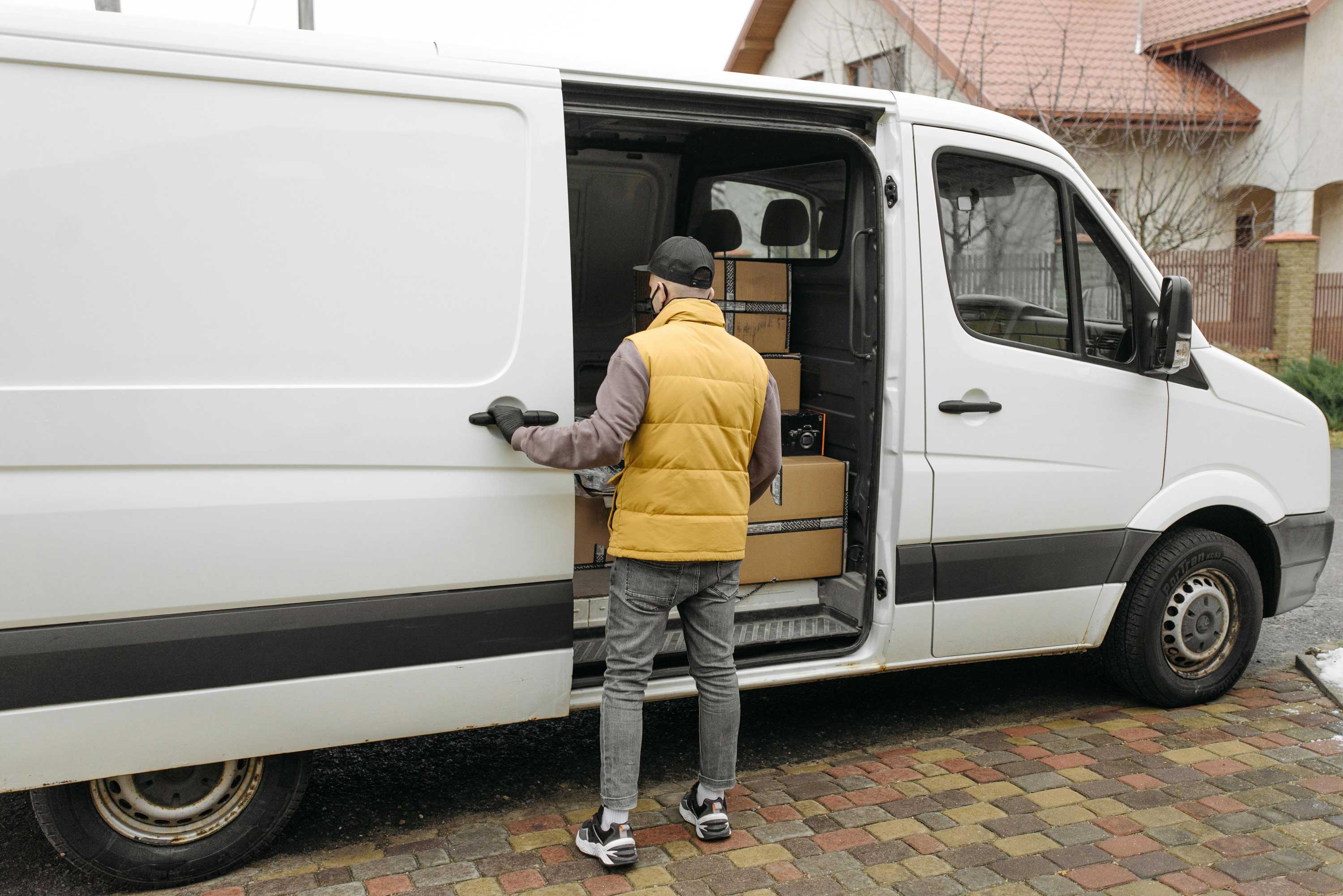 Man wearing a yellow vest and black cap loading cardboard boxes into the back of a white delivery van.