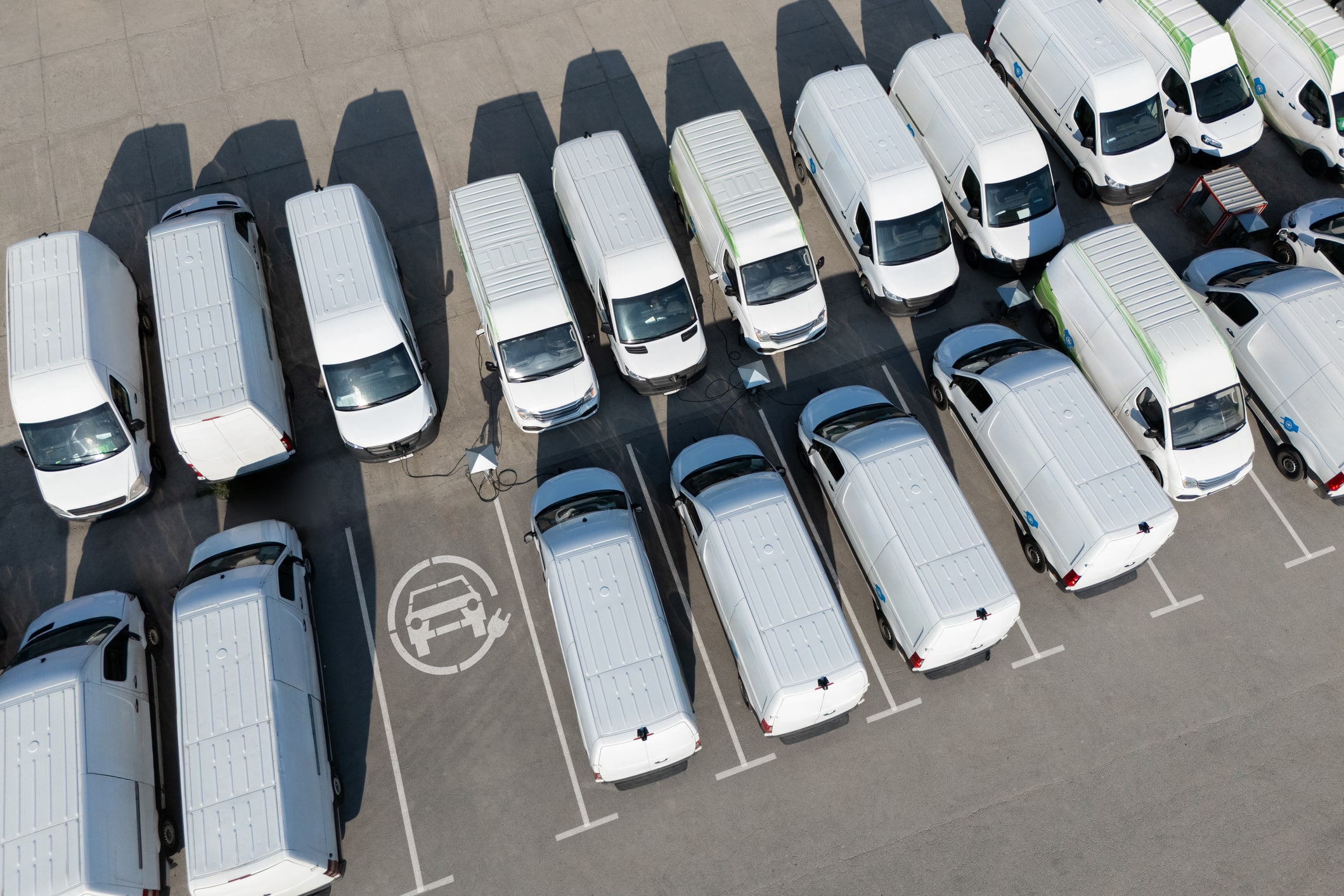 Aerial view of a parking lot with white electric vans charging at charging stations.