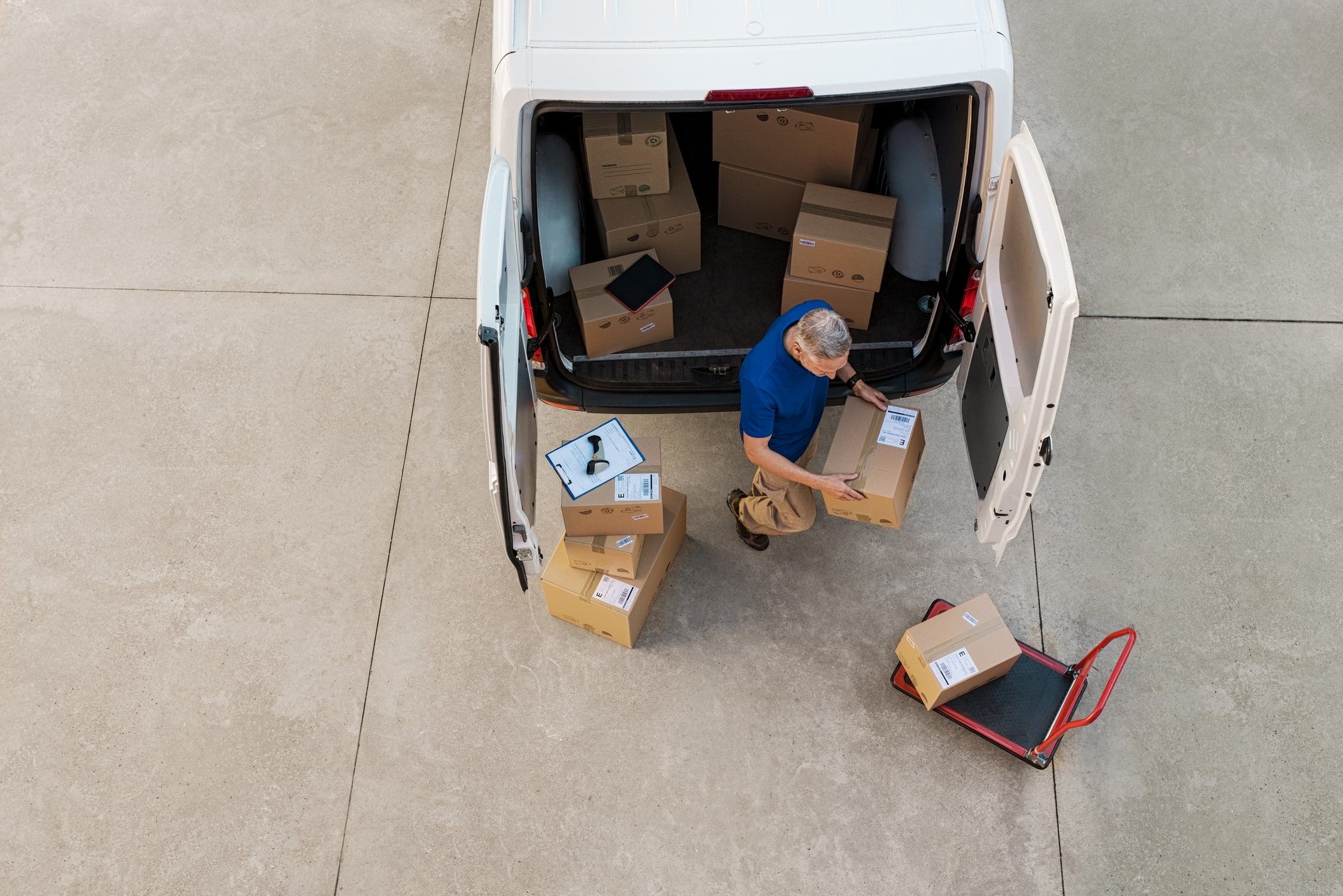 A delivery worker holding a cardboard box behind an open white van filled with packages, with more boxes and a clipboard nearby on the ground.
