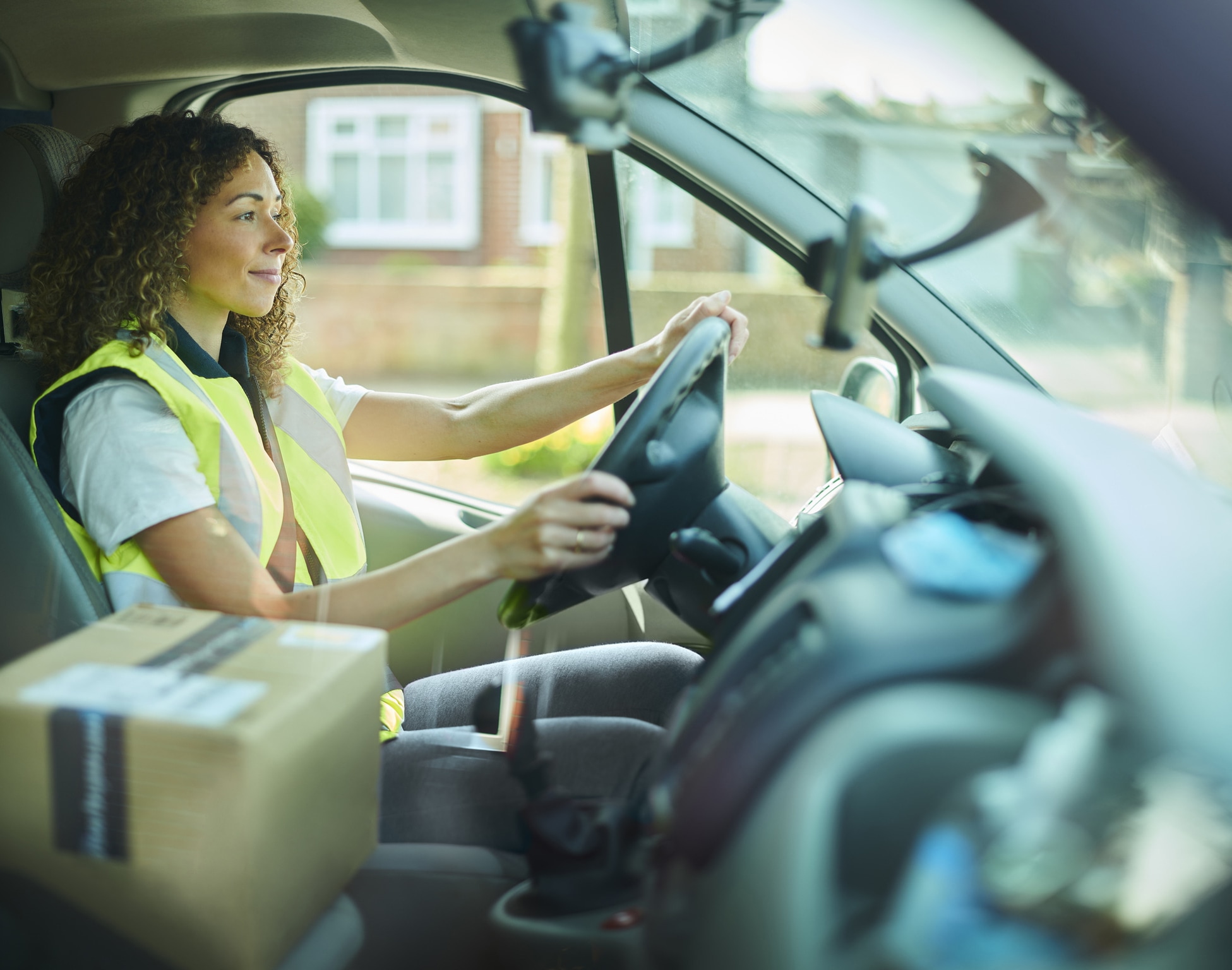 Woman wearing a high-visibility vest driving a delivery van with a package on the passenger seat.