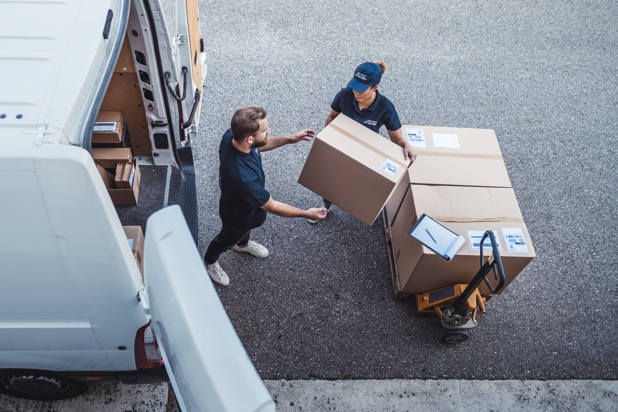 Two delivery workers unloading a large cardboard box from a white van onto a pallet jack stacked with more boxes and a clipboard.