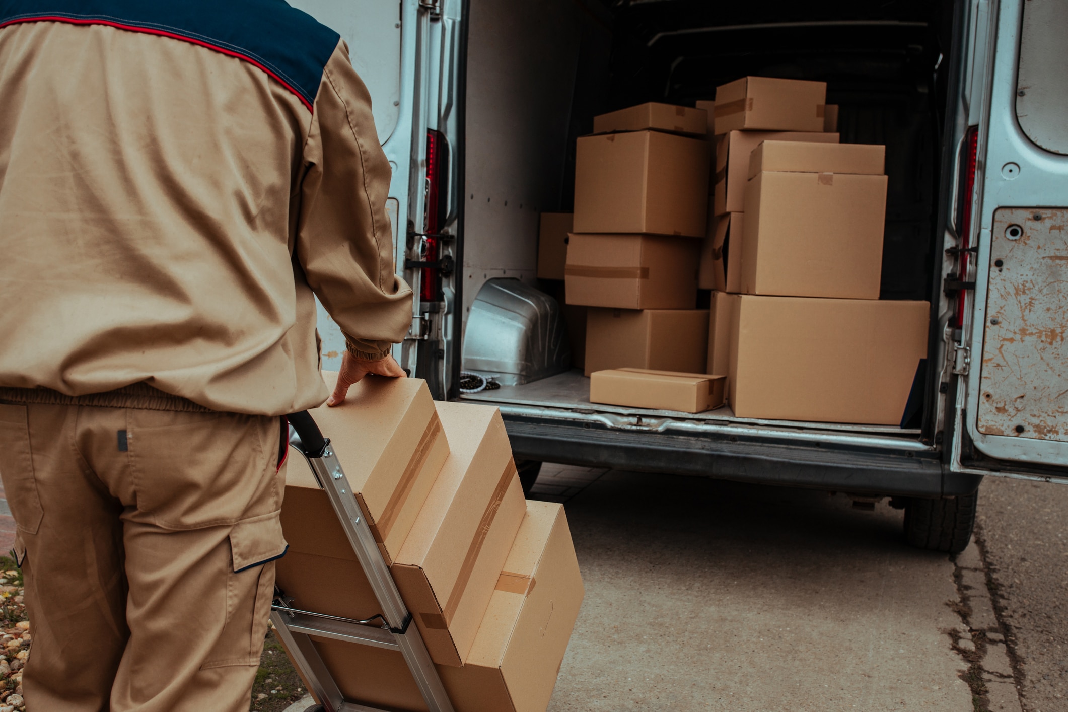 Delivery person unloading cardboard boxes from the back of a white van using a hand truck.