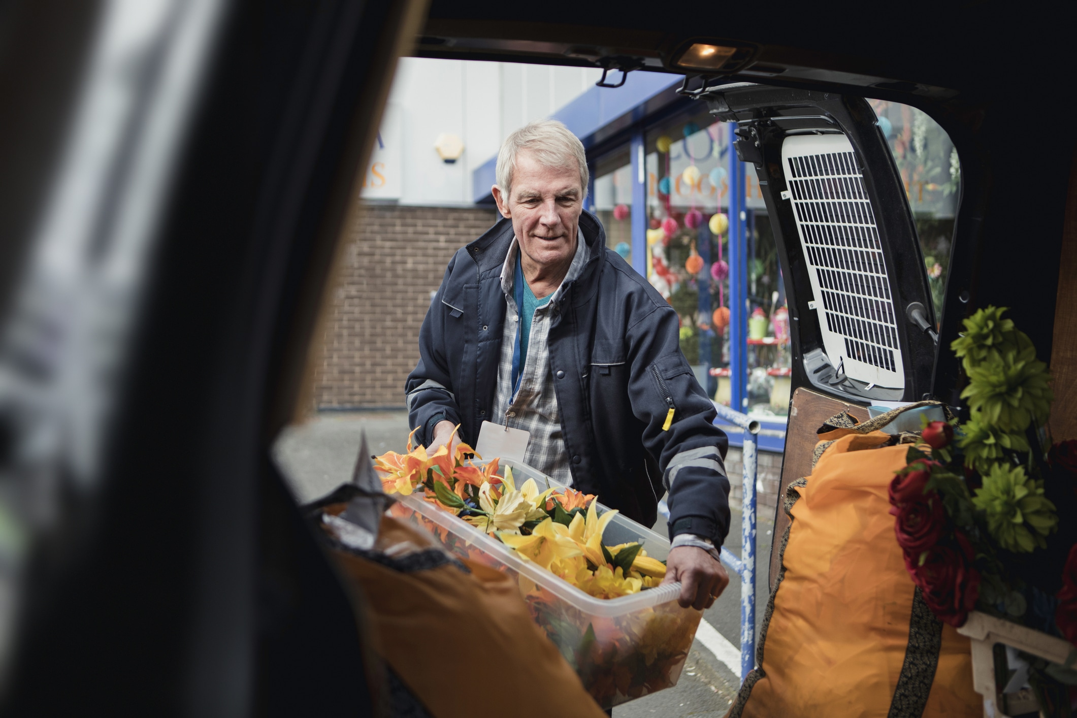 Older man unloading a plastic container filled with yellow and orange artificial flowers from the back of a vehicle near a shop window.