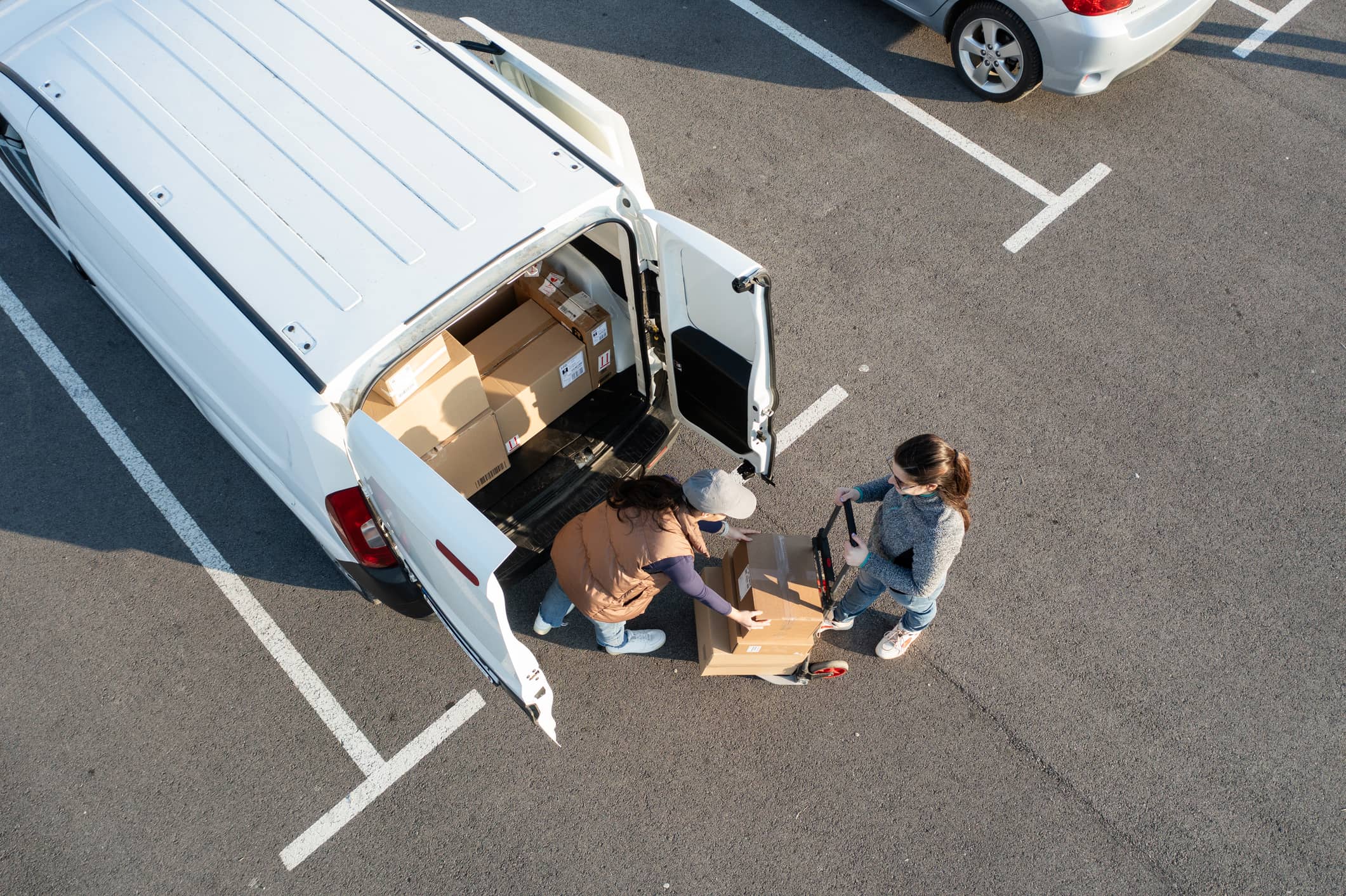 Two people unloading cardboard boxes from a white van in a parking lot, using a hand dolly.