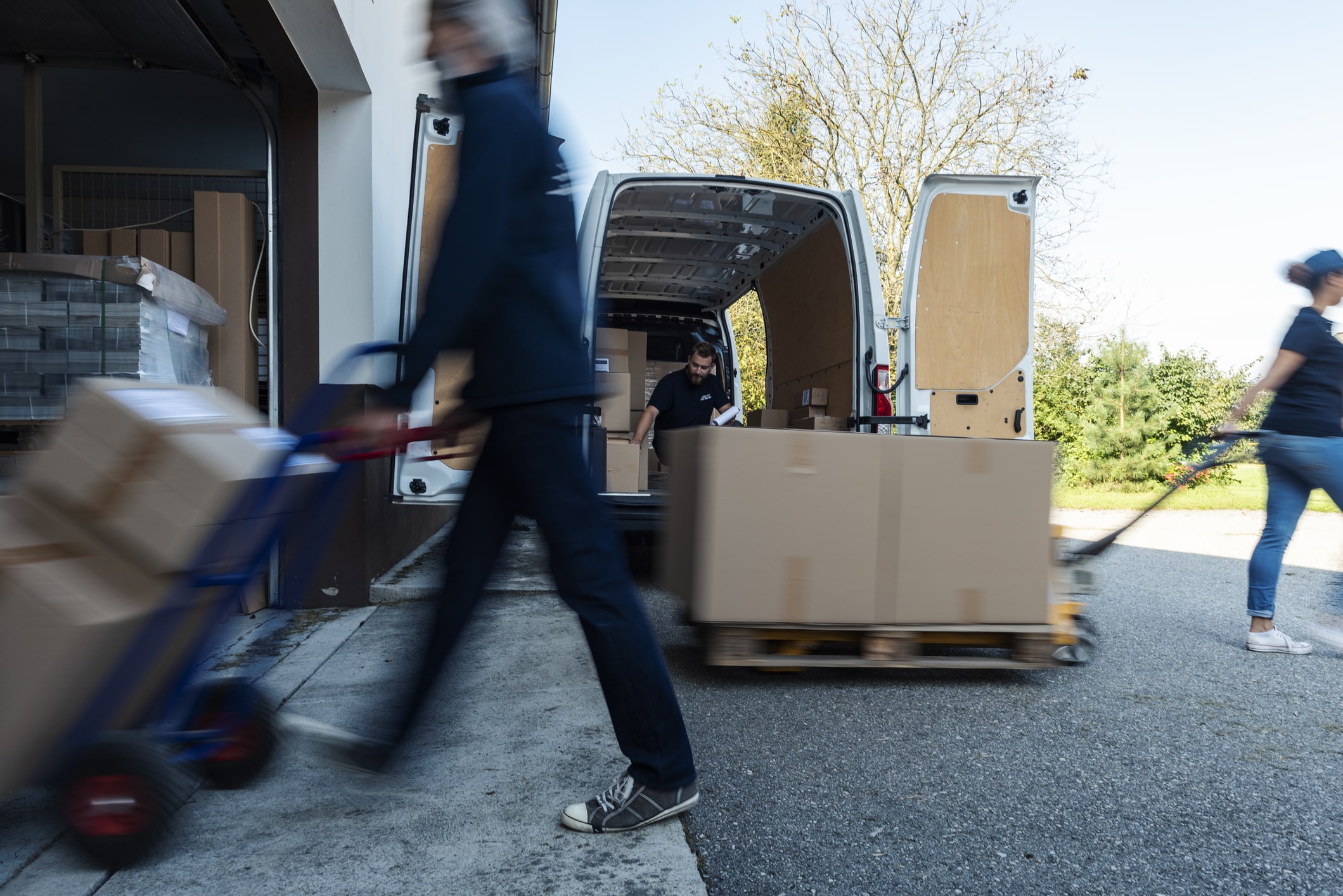 Workers moving cardboard boxes with hand trucks near a white delivery van with open rear doors.