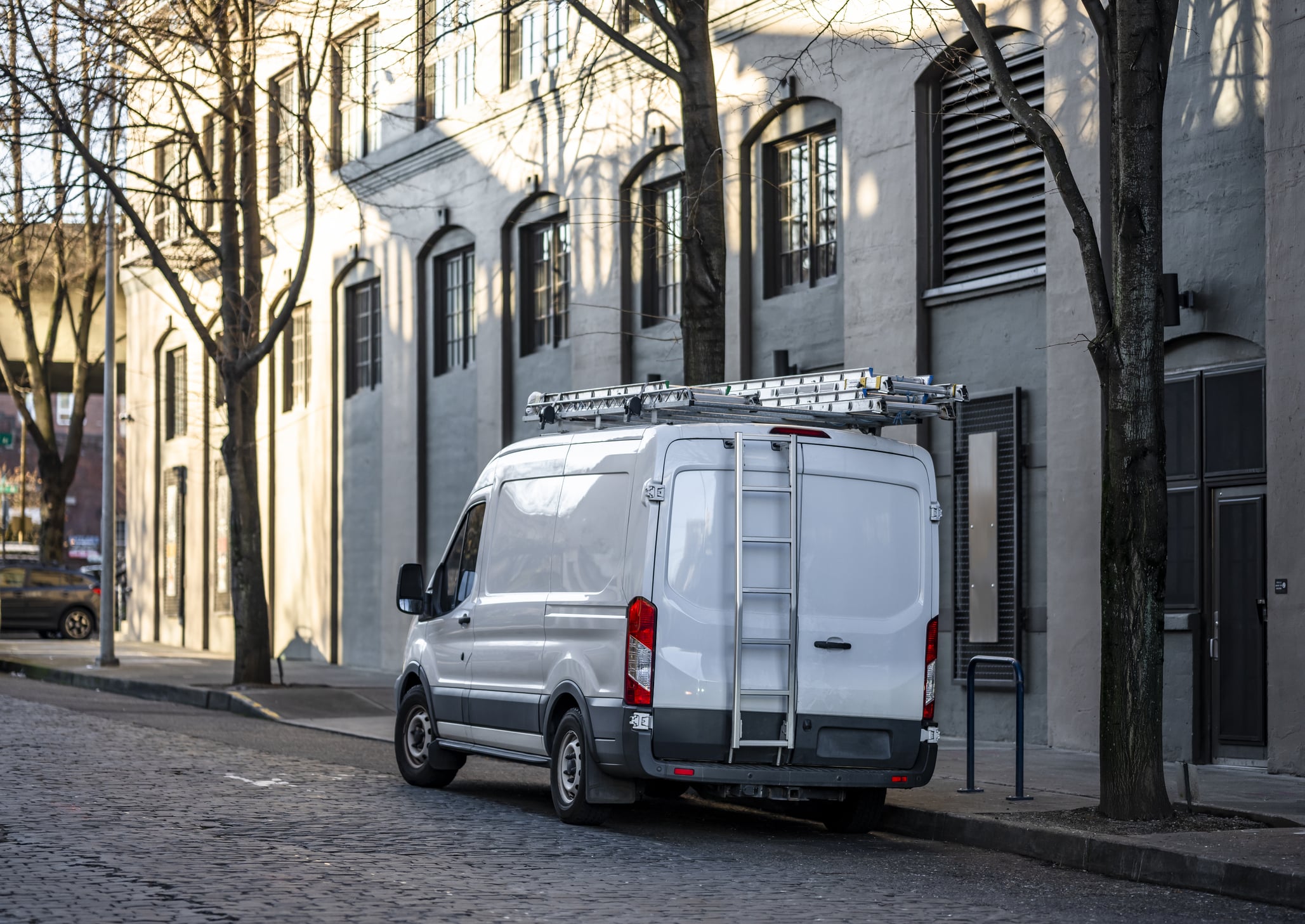 White cargo van with ladders mounted on the roof, parked on a cobblestone street next to a building.
