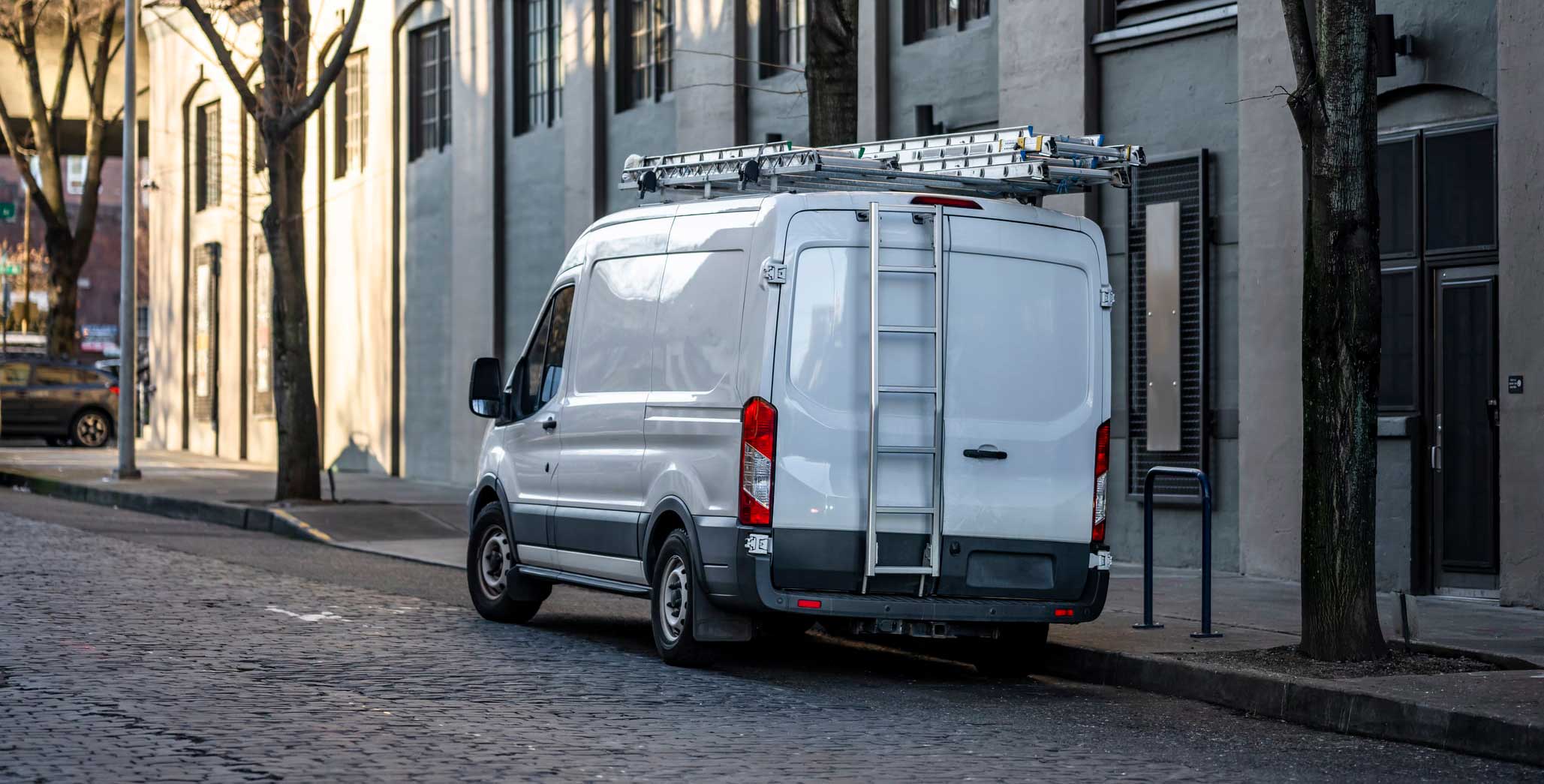 White cargo van with ladders mounted on the roof, parked on a cobblestone street next to a building.