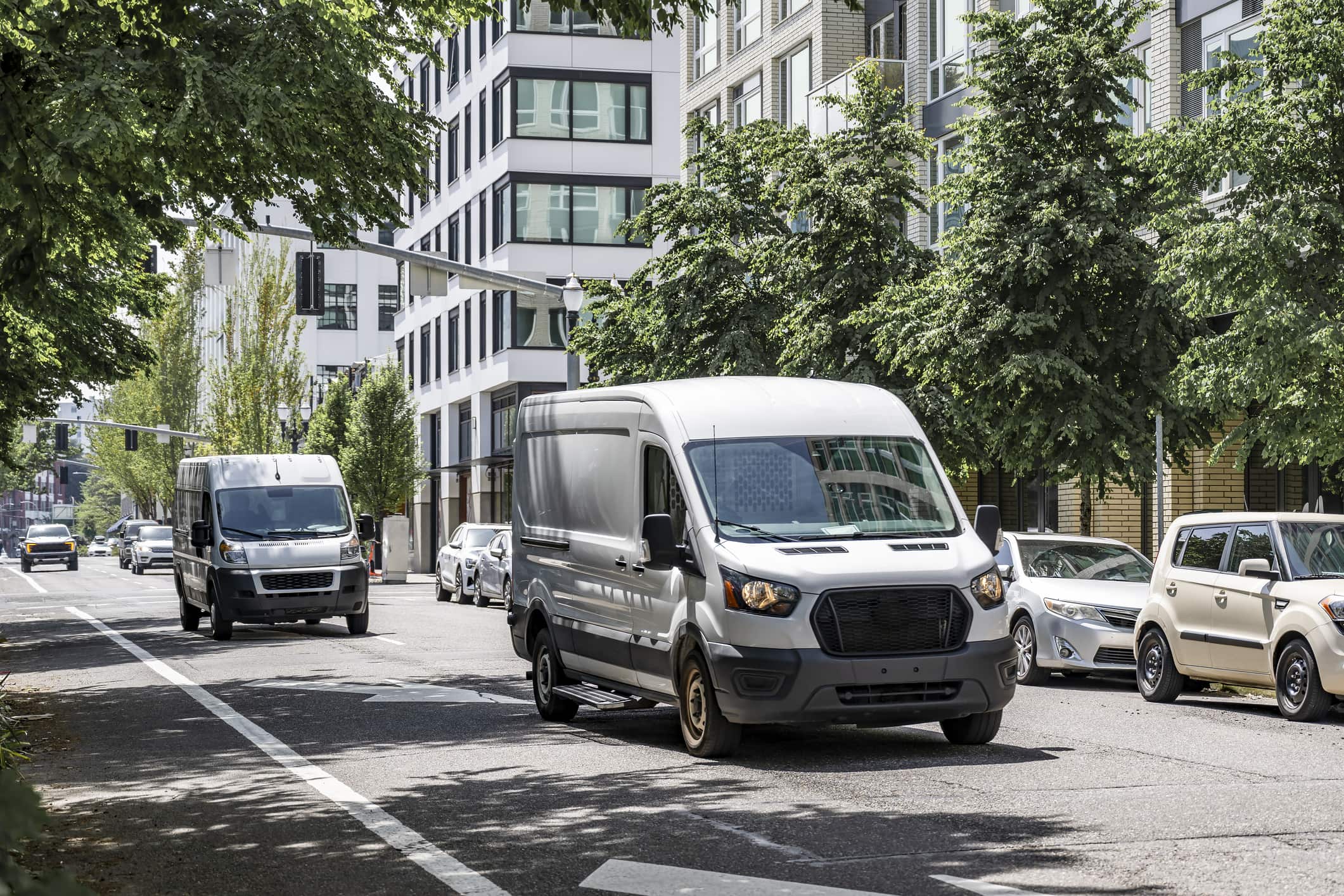 Two silver panel vans driving on a tree-lined city street with modern buildings and parked cars.