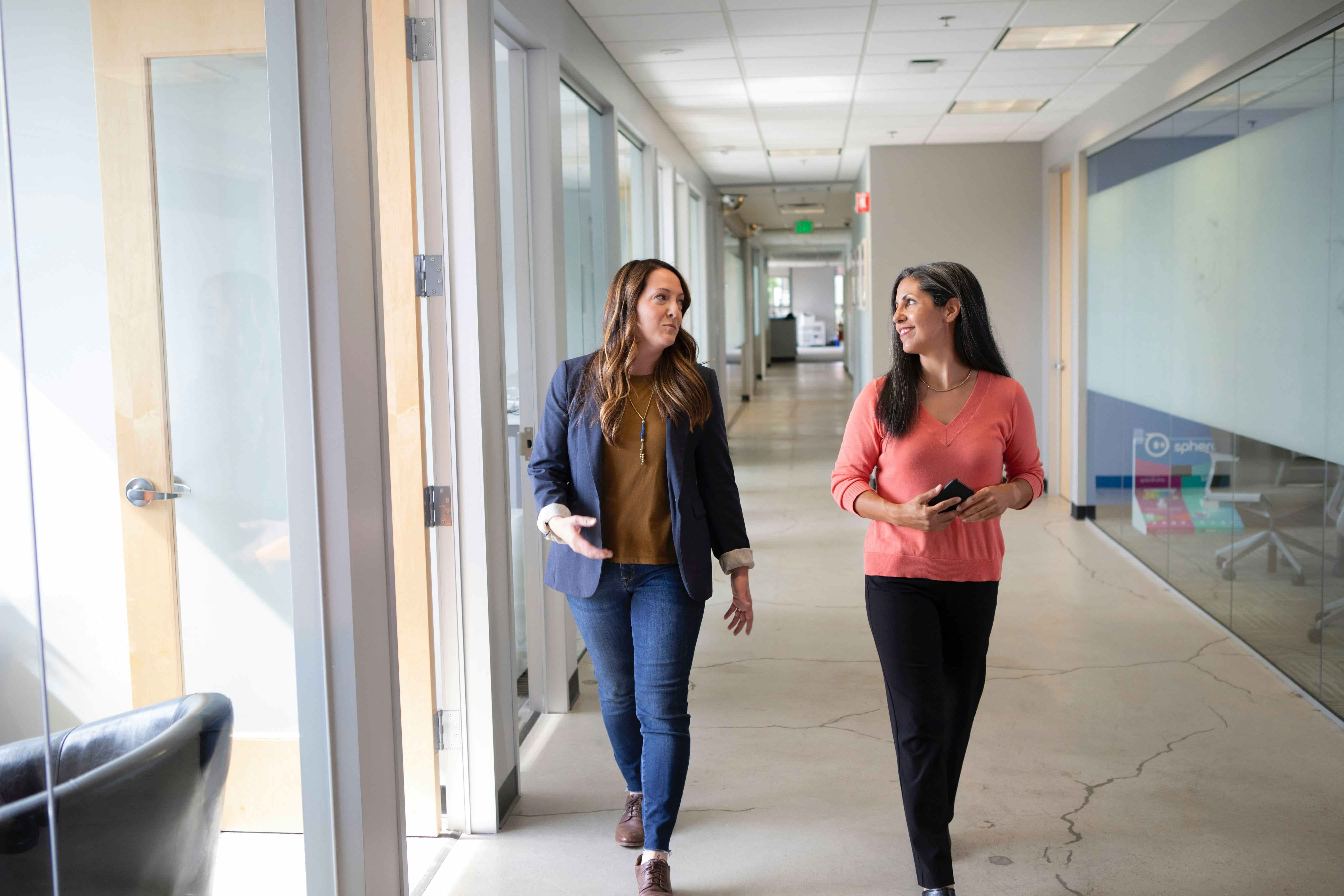 Two women walking and talking in a modern office hallway with glass walls and doors.