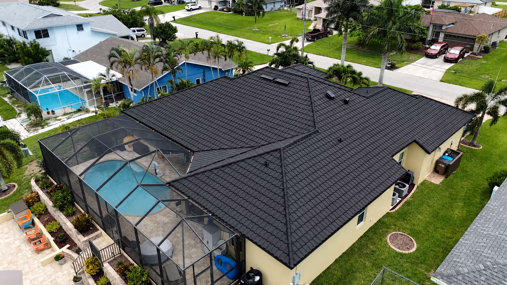 Aerial view of a beige house with a dark tiled roof and screened-in backyard pool in a green neighborhood.