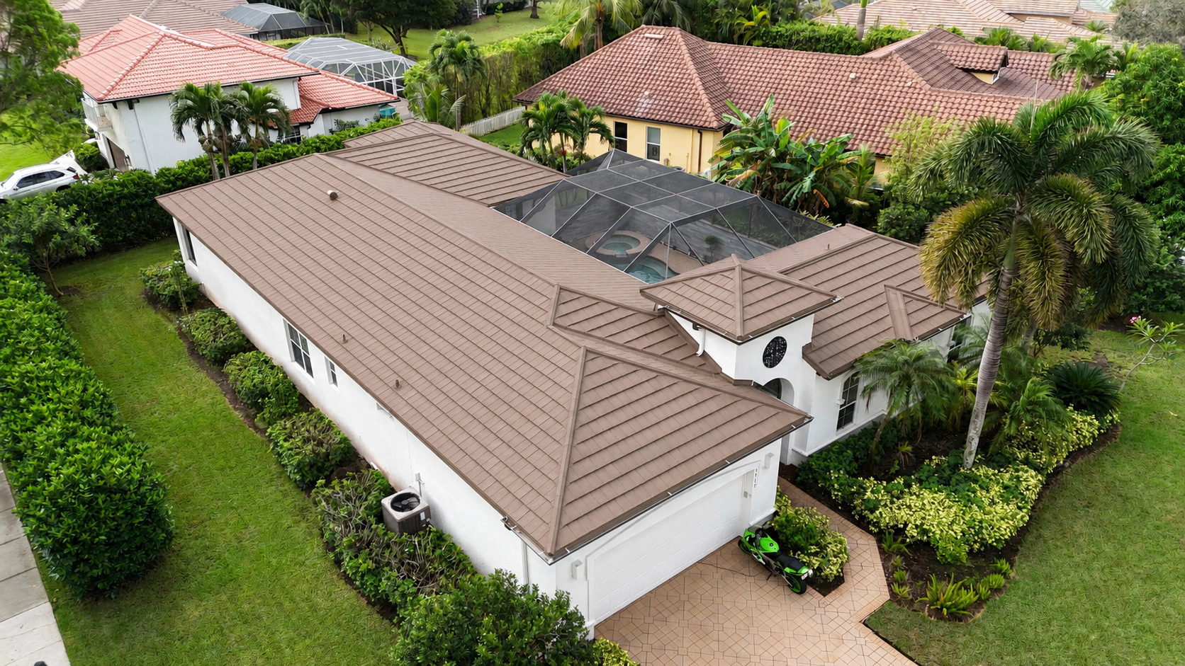 Aerial view of a white house with a brown tiled roof, palm trees, and a screened pool area.