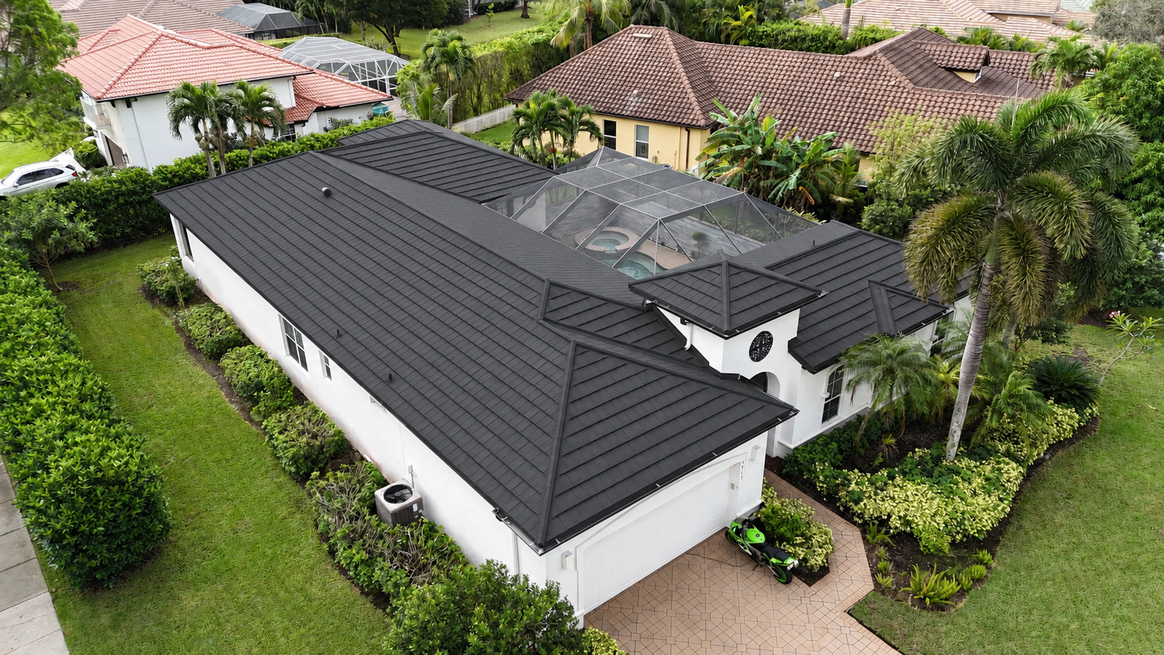 Aerial view of a white house with a black roof and screened pool enclosure surrounded by greenery.