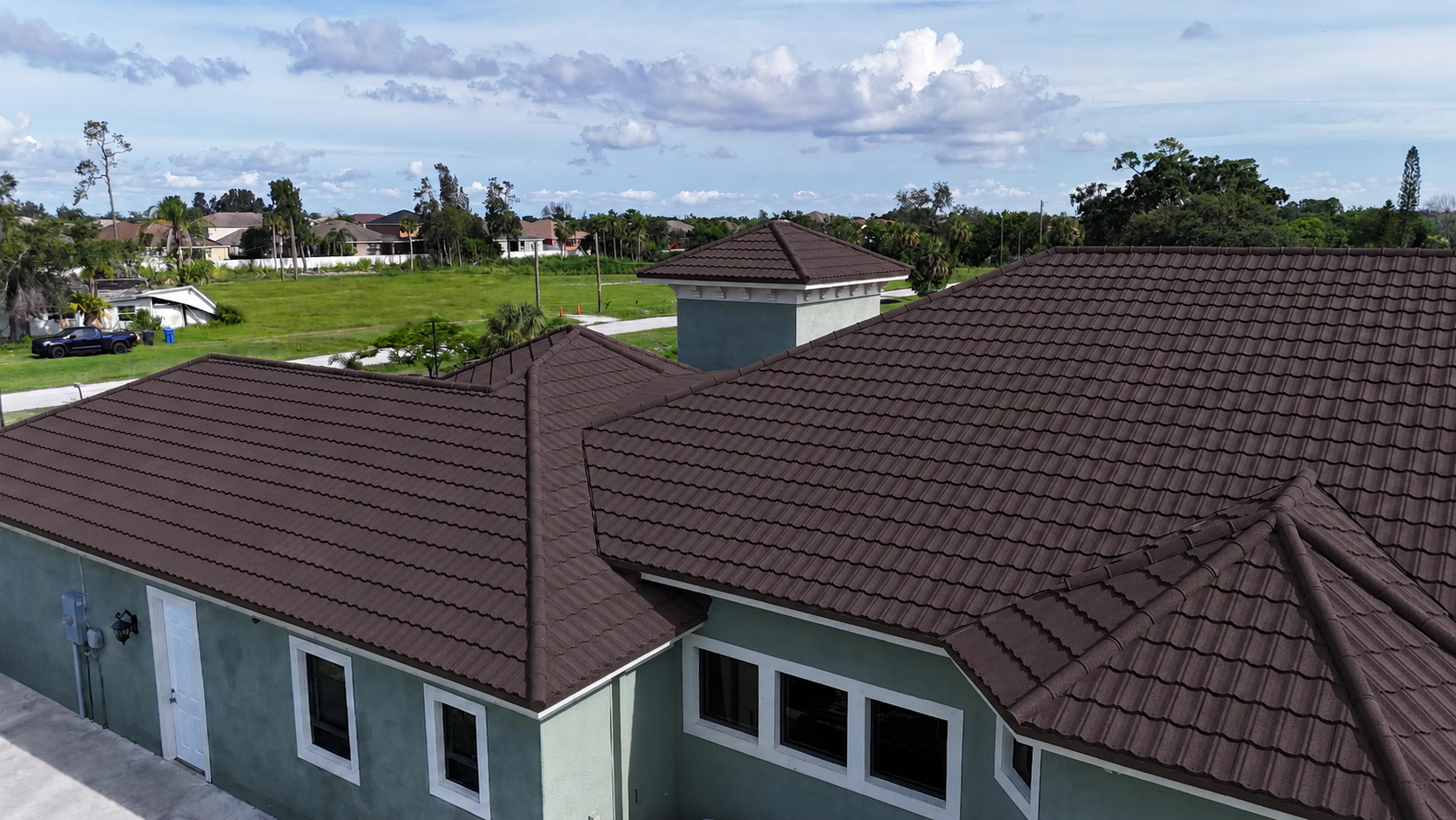 Residential house with multiple peaked brown tiled roofs under a partly cloudy blue sky.
