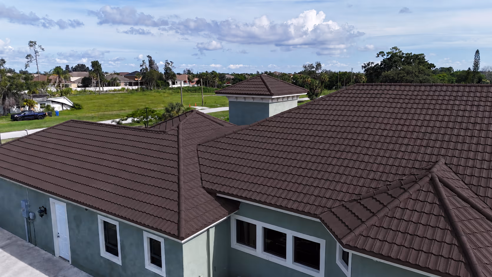 Residential house with multiple peaked brown tiled roofs under a partly cloudy blue sky.