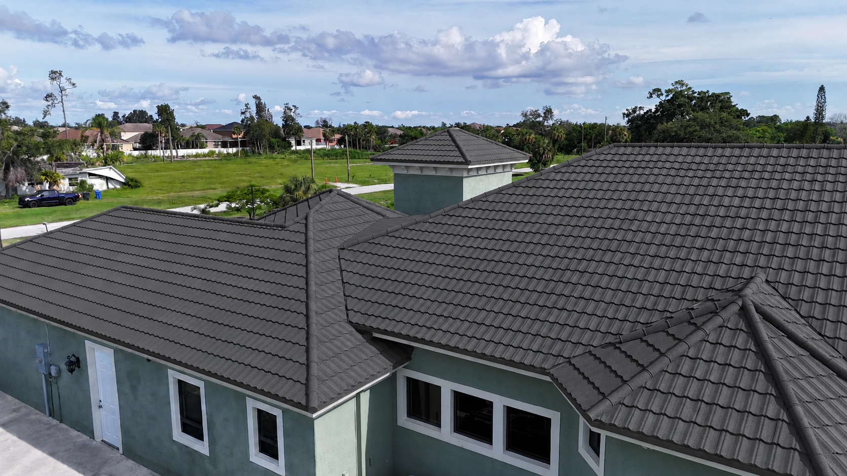 Grey tiled roof on a green house under a partly cloudy blue sky with trees in the background.