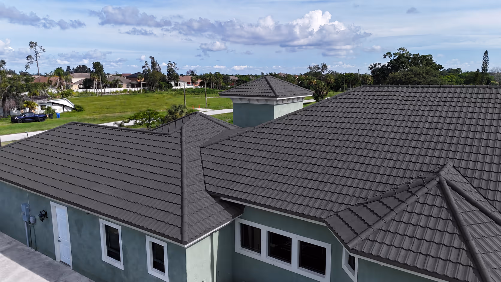 Grey tiled roof on a green house under a partly cloudy blue sky with trees in the background.