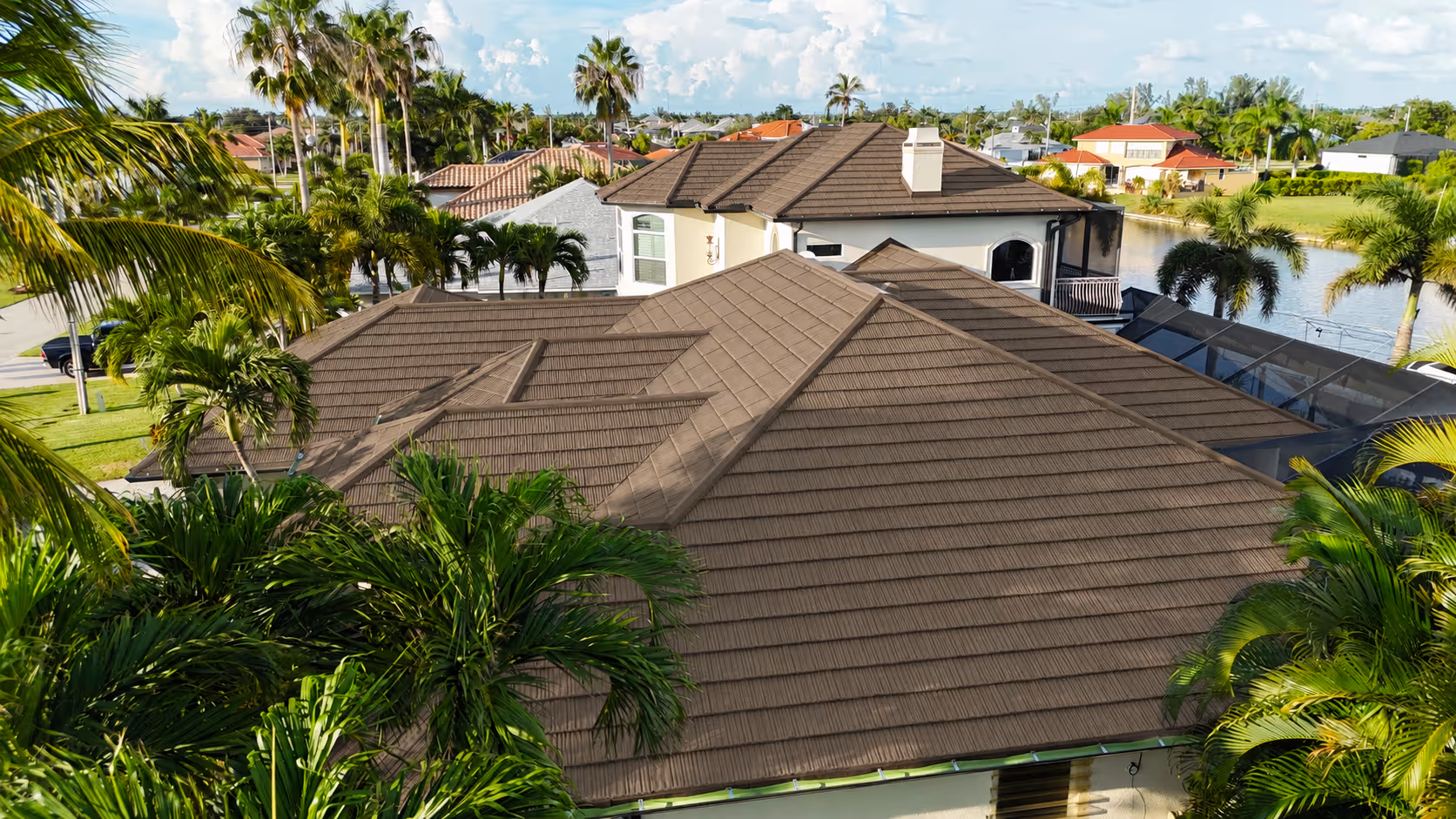 Aerial view of houses with brown tiled roofs surrounded by palm trees in a sunny neighborhood.