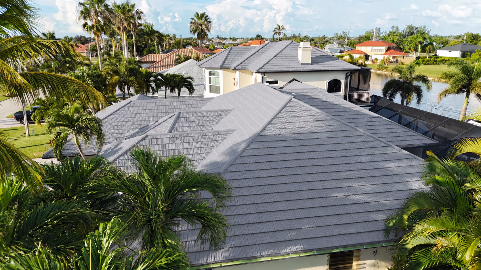 Aerial view of gray tile roofs on houses surrounded by palm trees and greenery in a sunny neighborhood.