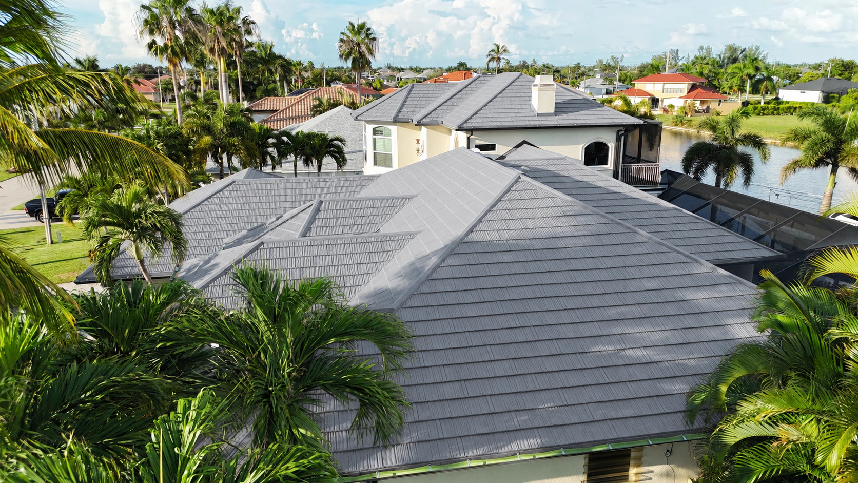 Aerial view of gray tile roofs on houses surrounded by palm trees and greenery in a sunny neighborhood.
