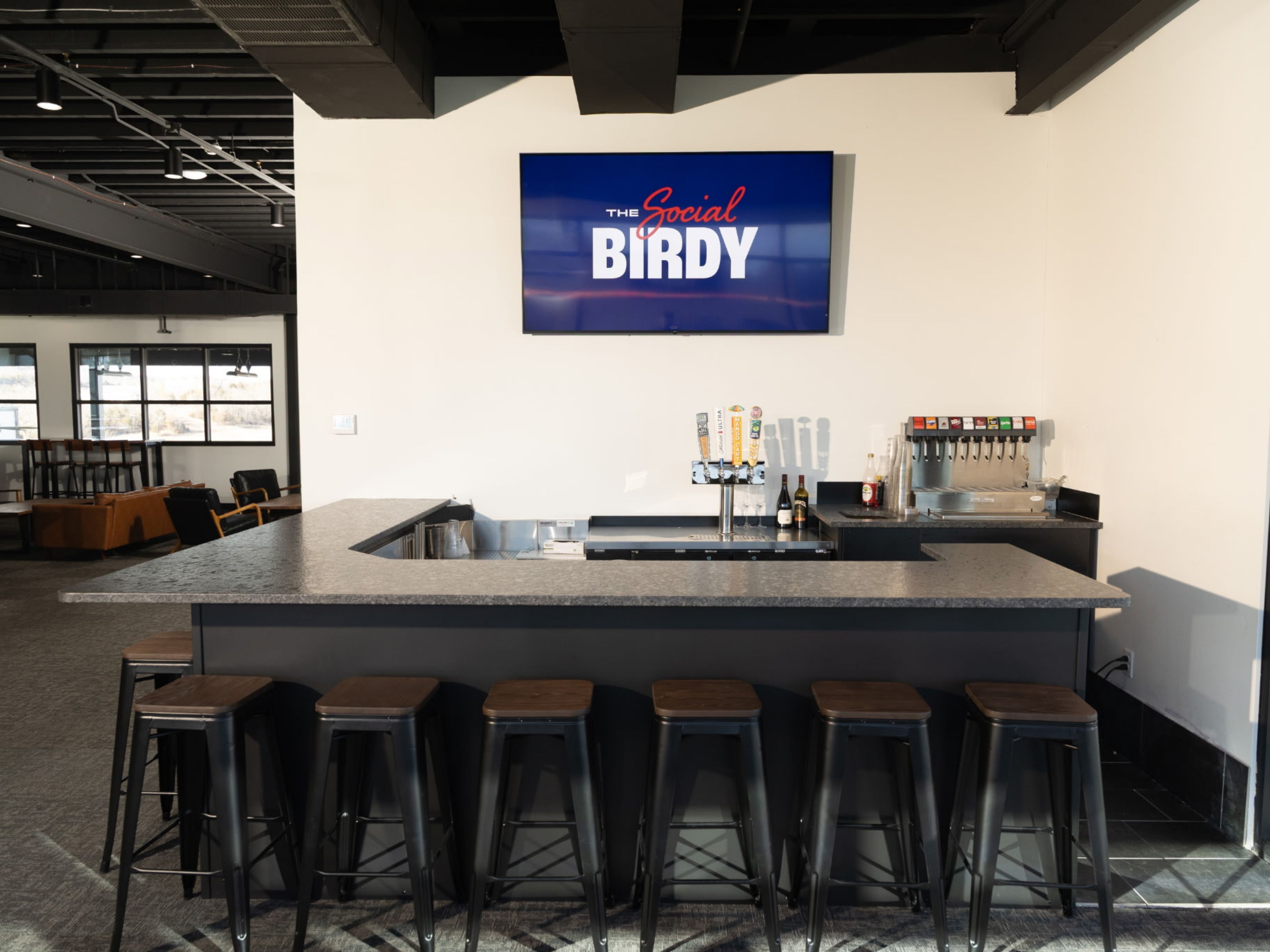 Modern bar counter with seven black metal stools, beer taps, and a soda dispenser under a screen displaying 'The Social Birdy'.