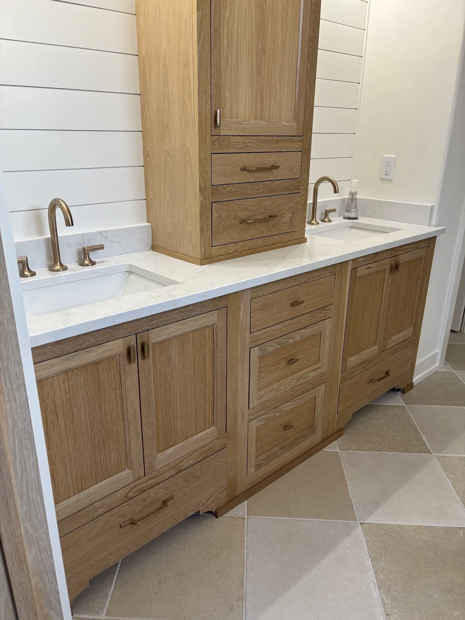 Modern bathroom vanity with double sinks, white marble countertops, light wood cabinetry, bronze faucets, and beige tiled floor.