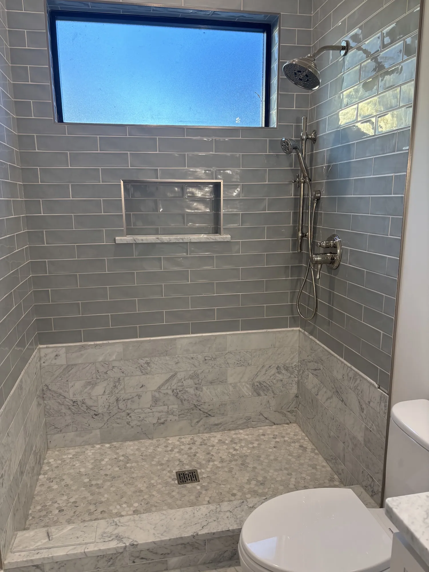 Modern walk-in shower with gray subway tiles and marble accents, featuring a built-in niche and stainless steel shower fixtures under a rectangular window.