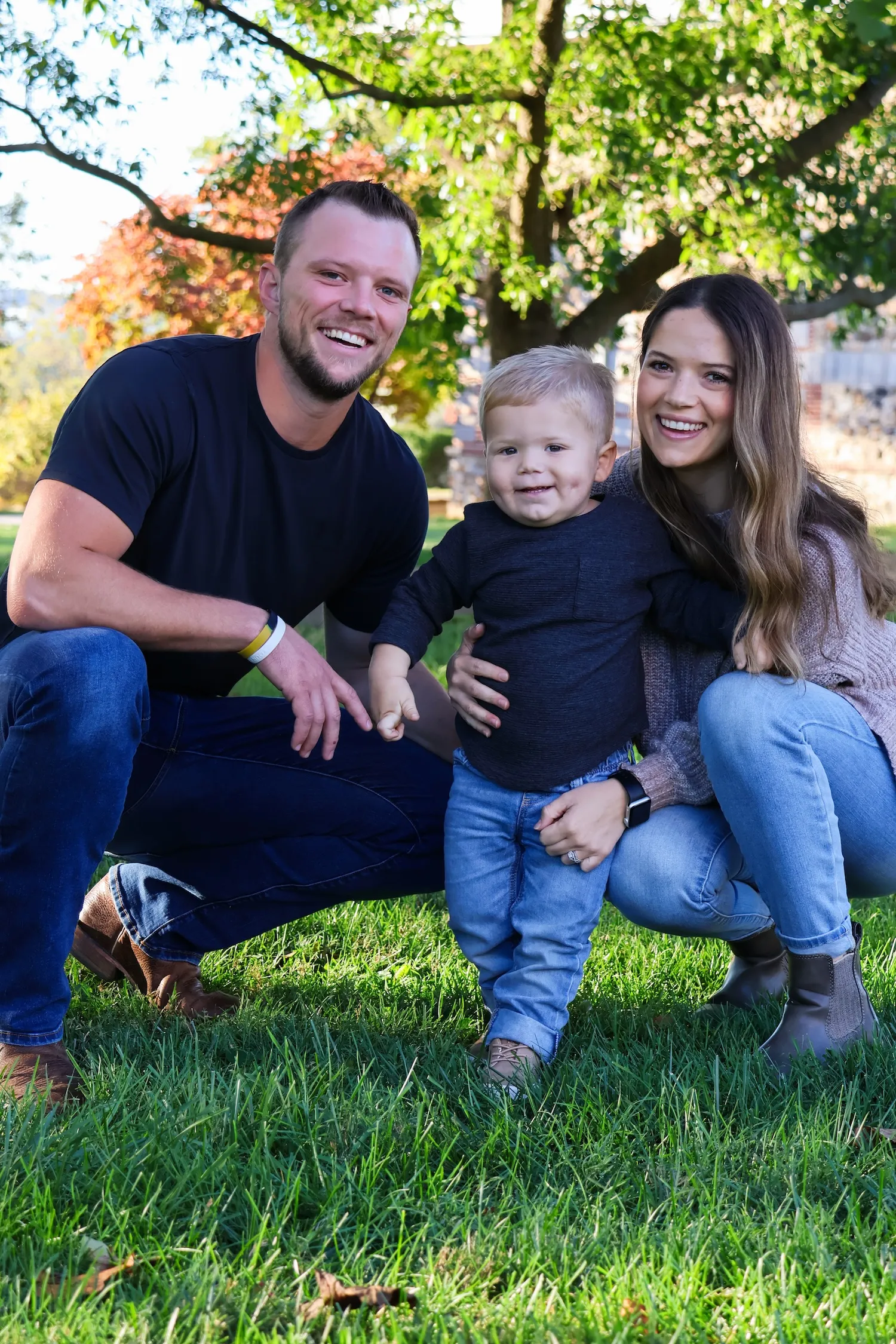 Oliver family crouching outdoors on grass with a toddler standing between them under a leafy tree.