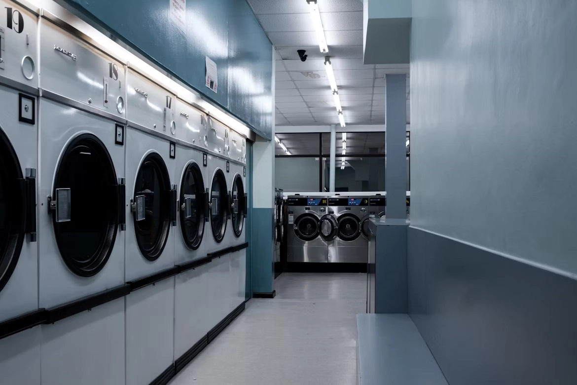 Interior of a laundromat with rows of front-loading washing machines and dryers along a light blue wall.
