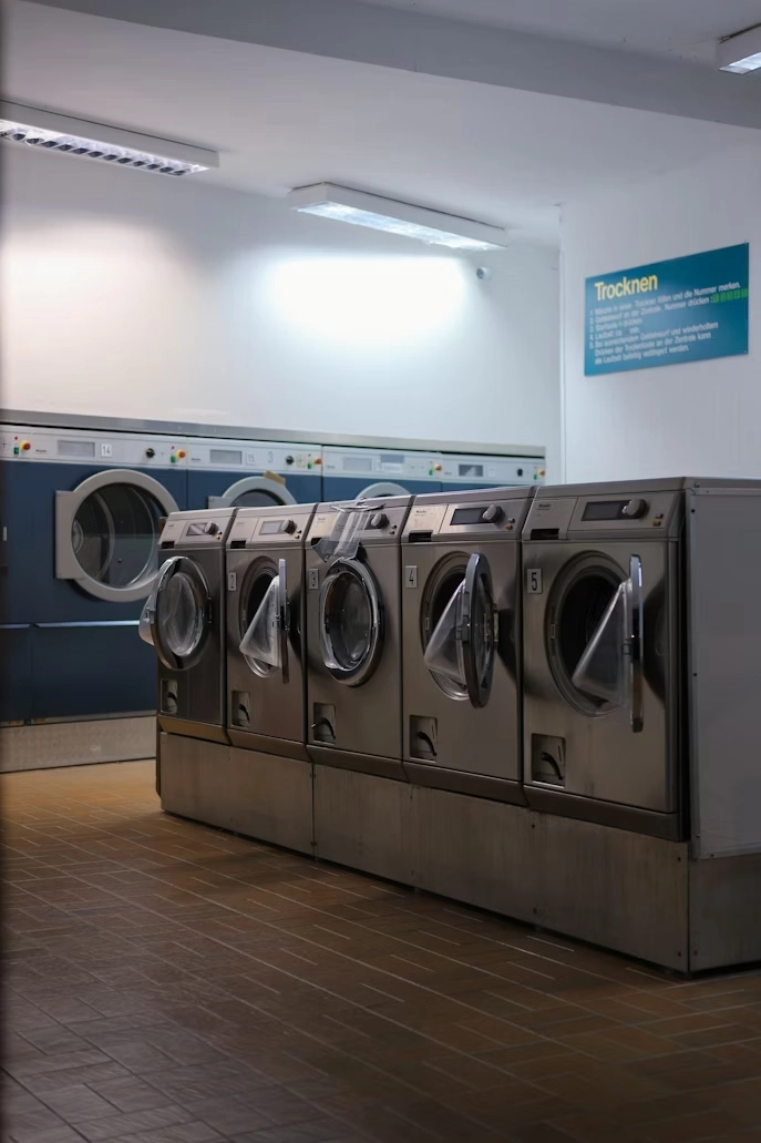 A dimly lit laundromat with rows of industrial washing machines and dryers. The flooring is brown tile, and a sign with text is on the wall.