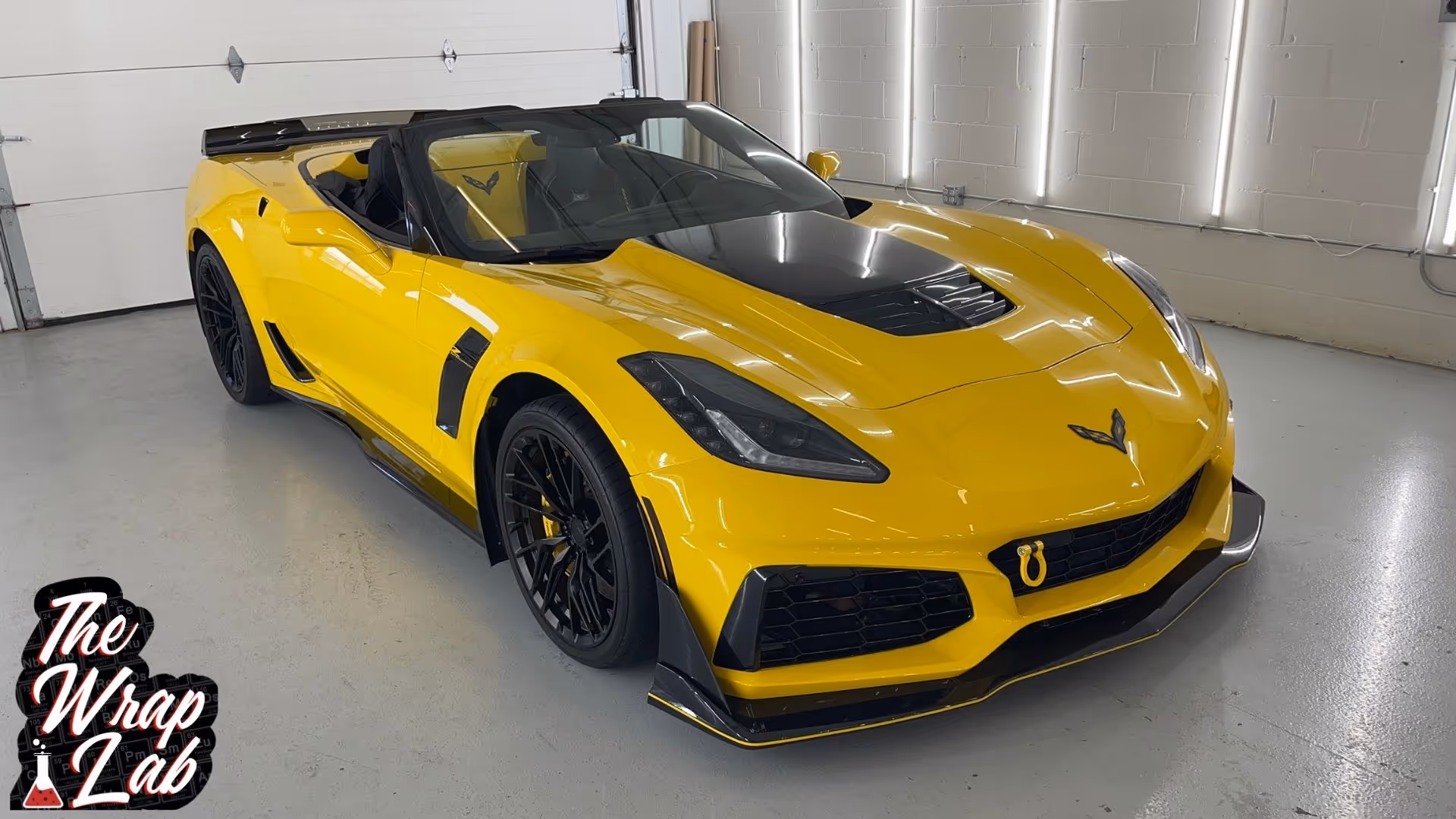 A bright yellow convertible sports car with a sleek design and black accents is parked in a well-lit garage. The car features a bold front grille, matte black hood, and stylish wheels, showcasing its premium appearance from The Wrap Lab.