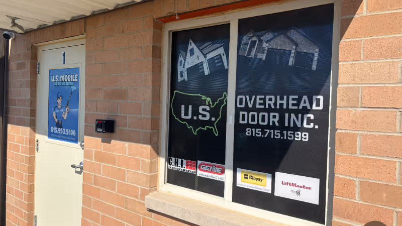 An exterior view of a brick building showing two windows. One displays the logo for "U.S. Mobile" with a phone number. The other features a sign for "U.S. Overhead Door Inc." including a phone number and various brands. The scene is bright and clear.