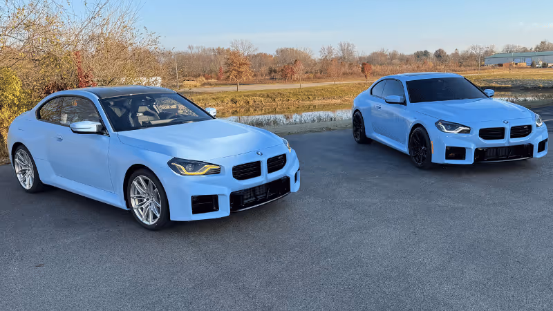 Two light blue BMW cars parked side by side on a paved surface, with a background of trees and grass. The vehicles display sleek designs, emphasizing their sporty features. The setting suggests a peaceful outdoor location, showcasing the Premium quality of The Wrap Lab.