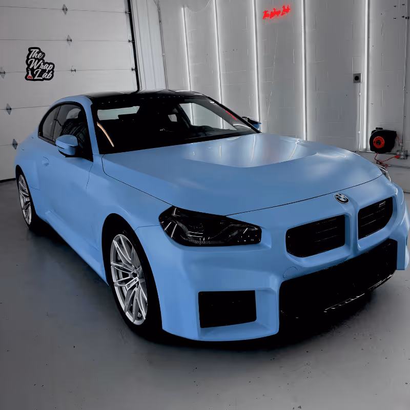 A light blue BMW coupe parked in a sleek garage with modern lighting. The car features an aggressive design with bold front grill and sporty wheels. The setting includes branding from The Wrap Lab on the wall.