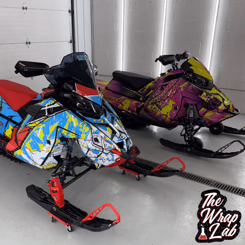 Two custom snowmobiles stand side by side in a well-lit garage. The first features a vibrant blue and red design with abstract patterns, while the second showcases a bold purple and yellow scheme, both equipped with black skis.