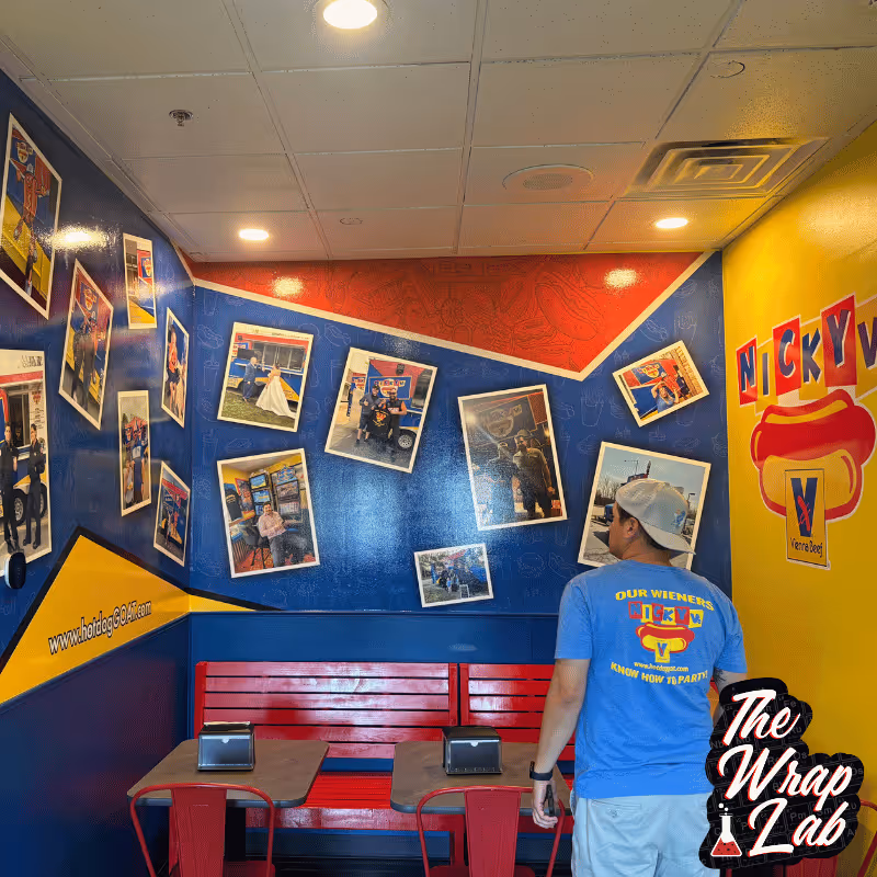 A man in a blue t-shirt stands in a colorful dining area filled with photographs on the walls, showcasing food and events. Bright blue and yellow paint creates a vibrant backdrop, complemented by red seating. The Wrap Lab Premium branding is visible.