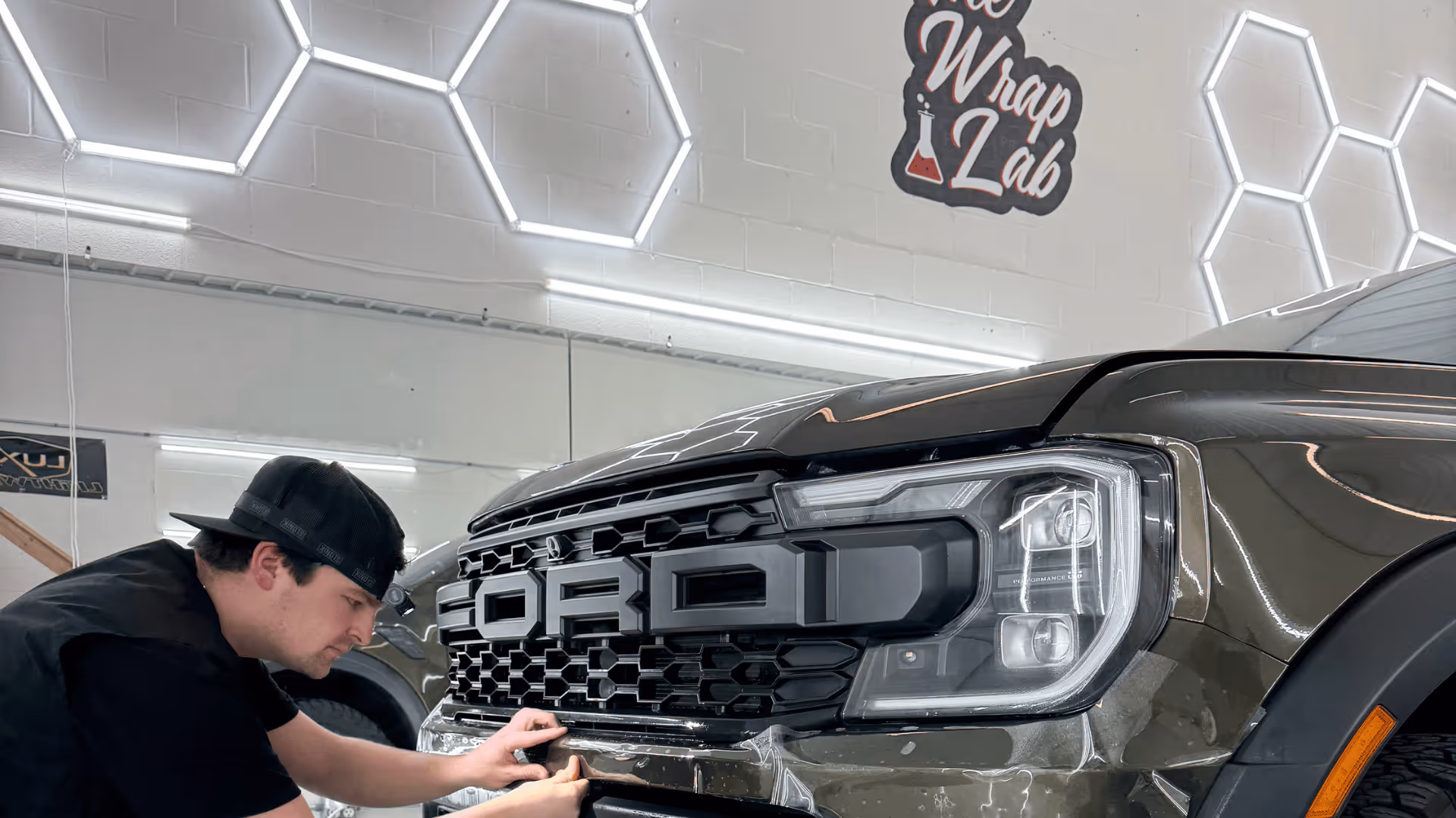 A technician applying a vinyl wrap to the front grille of a Ford vehicle in a modern shop. The backdrop features hexagonal lighting and a sign that reads 'The Wrap Lab Premium,' enhancing the professional atmosphere of the workspace.