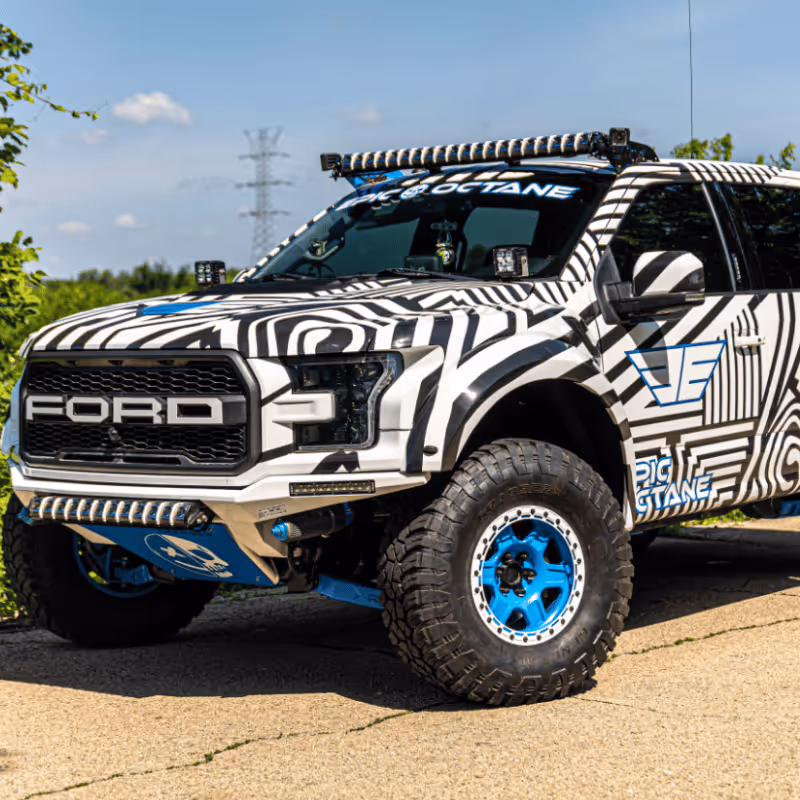 A customized Ford truck featuring a striking black and white zebra-striped wrap, accented with vibrant blue details. The vehicle is parked in a sunny outdoor setting, showcasing its rugged tires and off-roading features. The Wrap Lab Premium branding is visible.